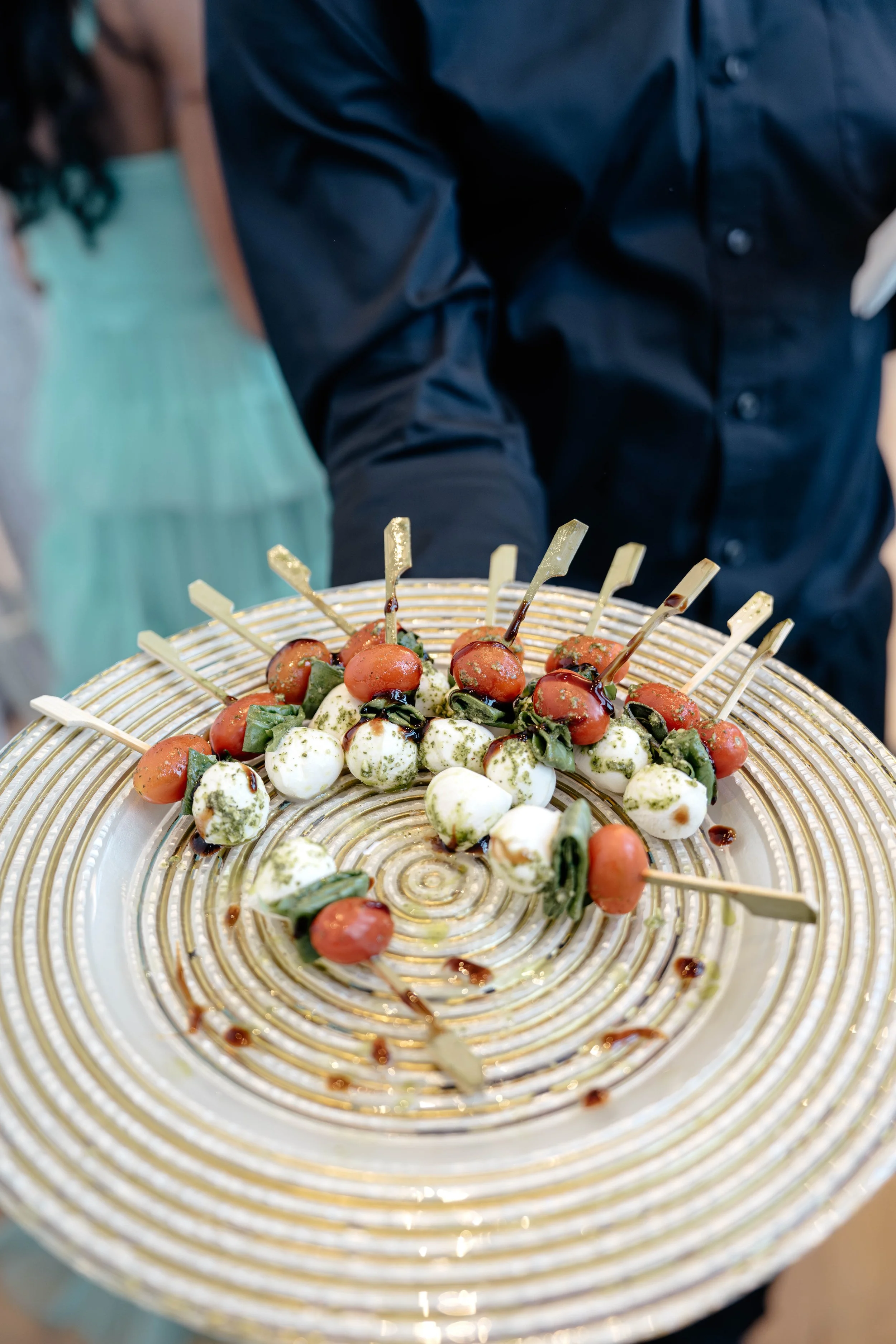 Skewers of mozzarella balls, cherry tomatoes, fresh basil, drizzled with balsamic glaze on a decorative gold-rimmed plate.