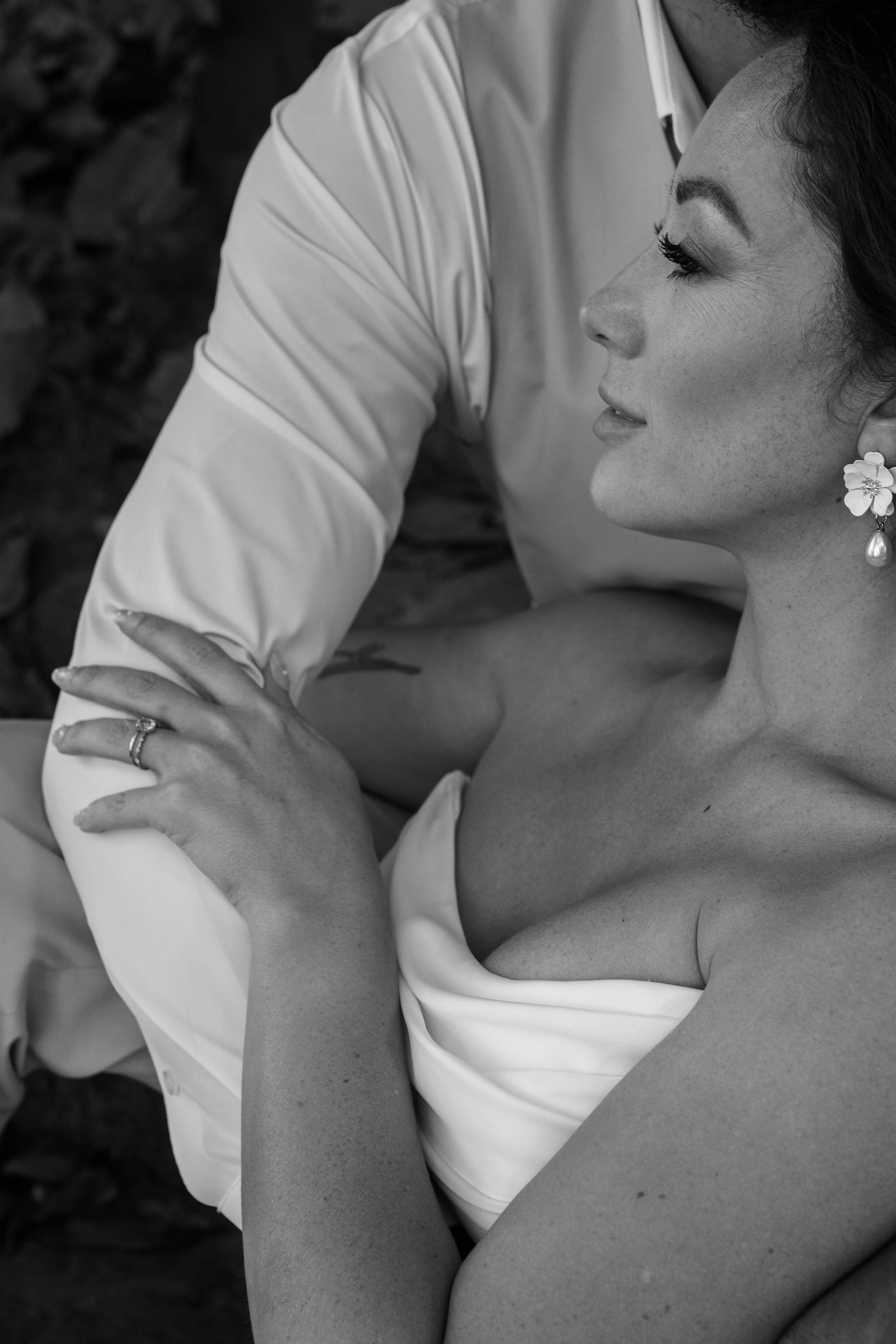 A black-and-white close-up of a woman with a floral earring and a tattoo, looking content, holding hands with a person wearing a white shirt, in an outdoor setting.