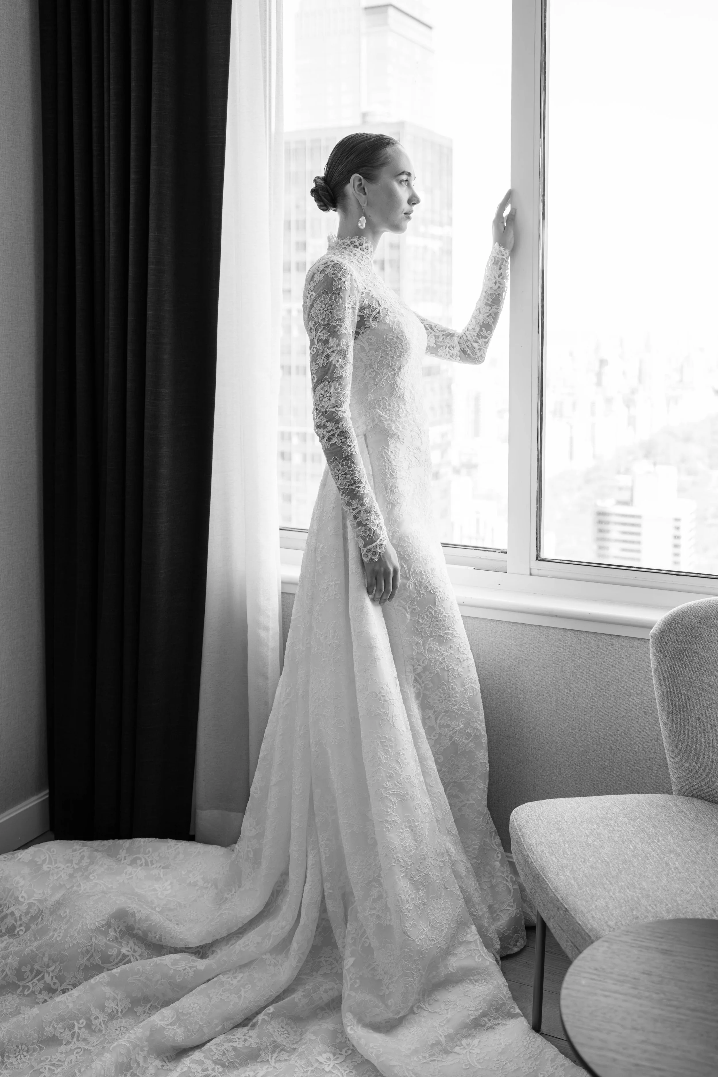 Black and white photograph of a woman in a lace wedding dress standing by a window, looking outside with a contemplative expression. The dress has a train that pools on the floor, and her hair is styled in a bun. A dark curtain partially covers the w
