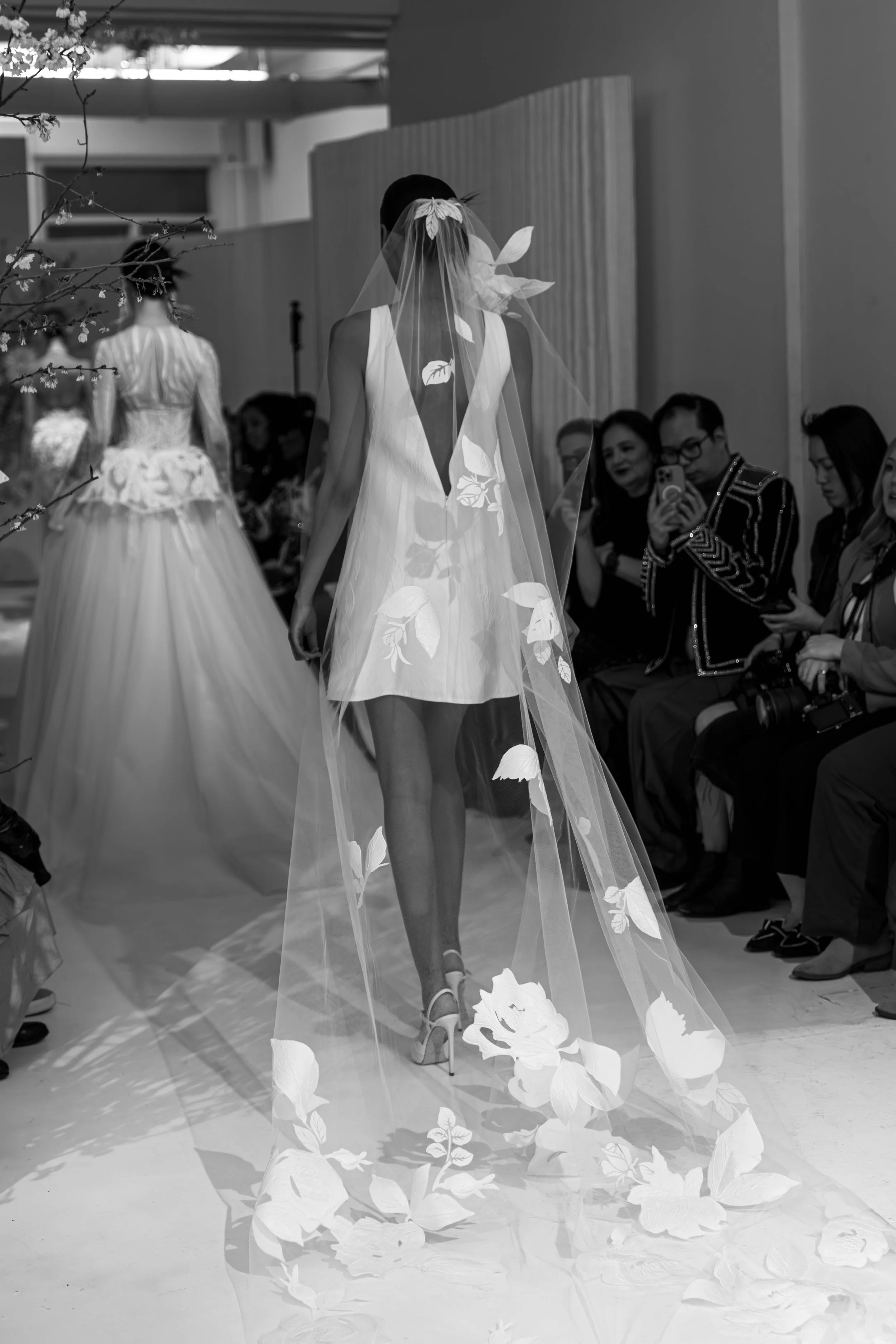 Black and white photo of a runway at a fashion show. A model is walking away from the camera, wearing a flowered dress and a large veil with leaves. Audience members are seated on either side, some taking photos, with the background showing various s
