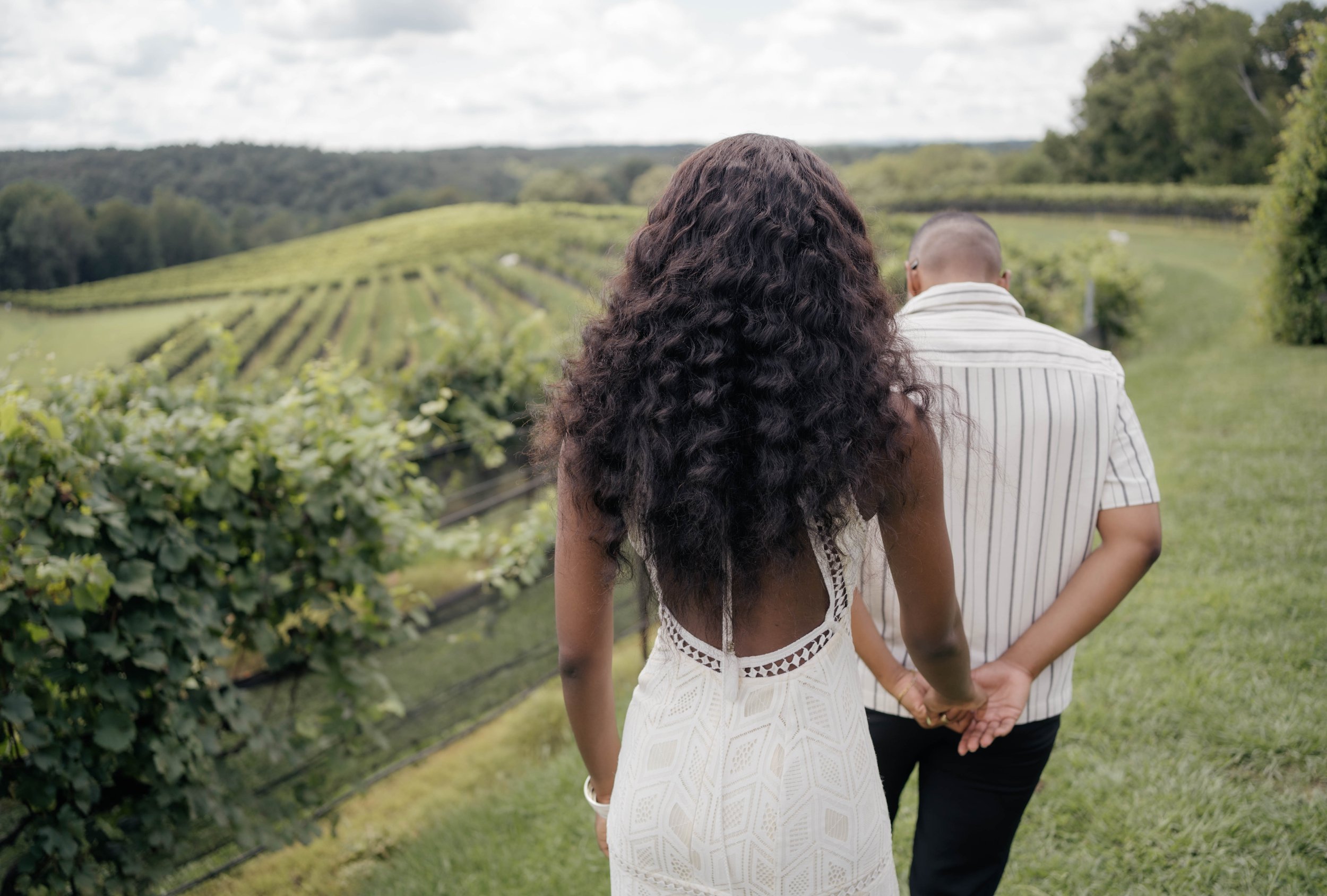 A couple holding hands and walking through a vineyard with rolling hills and trees in the background.