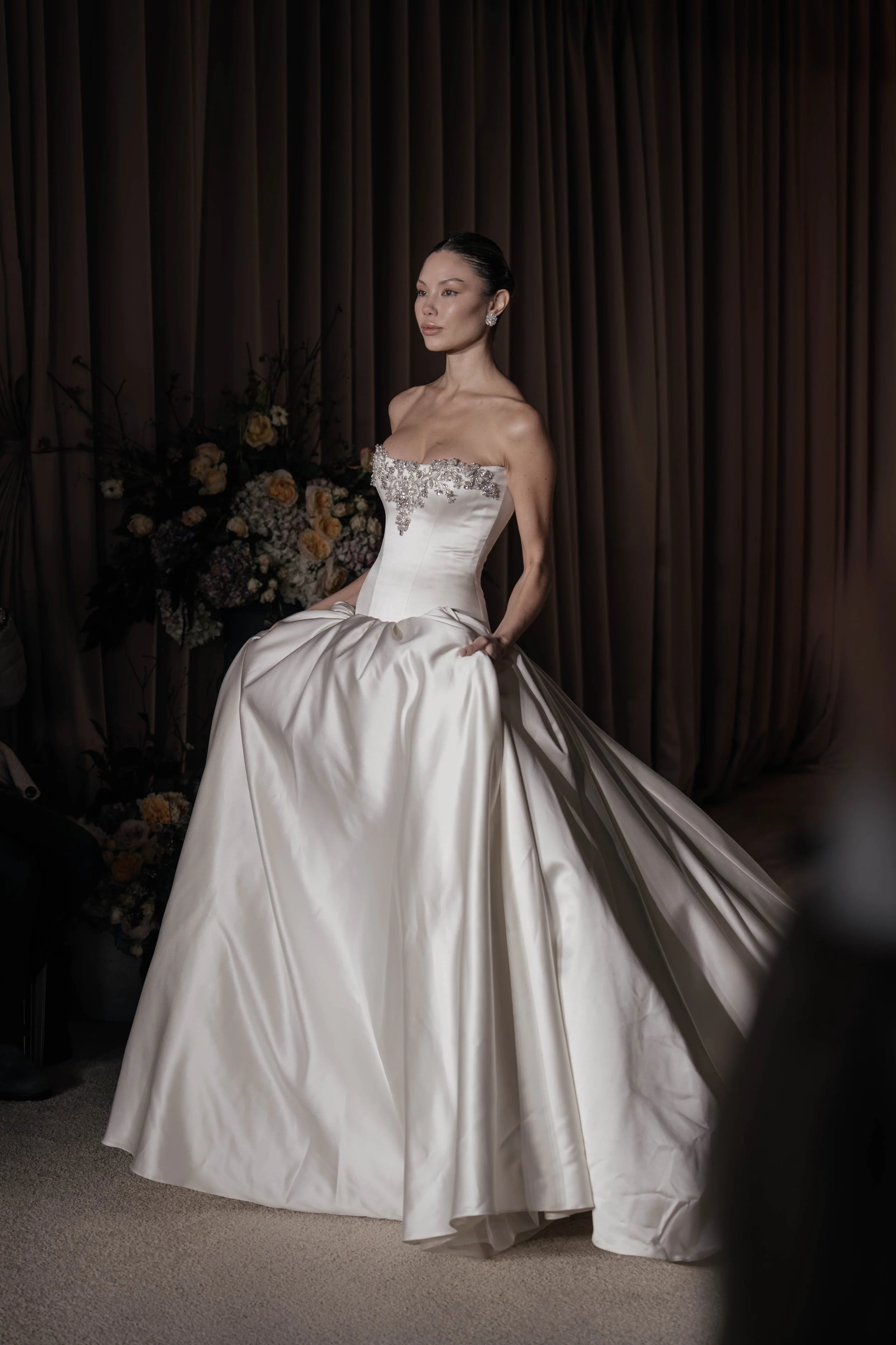 A woman in a white wedding gown with intricate beading on the bodice, sitting with one knee up. She has dark hair pulled back, earrings, and makeup. Behind her is a floral arrangement and brown curtains.