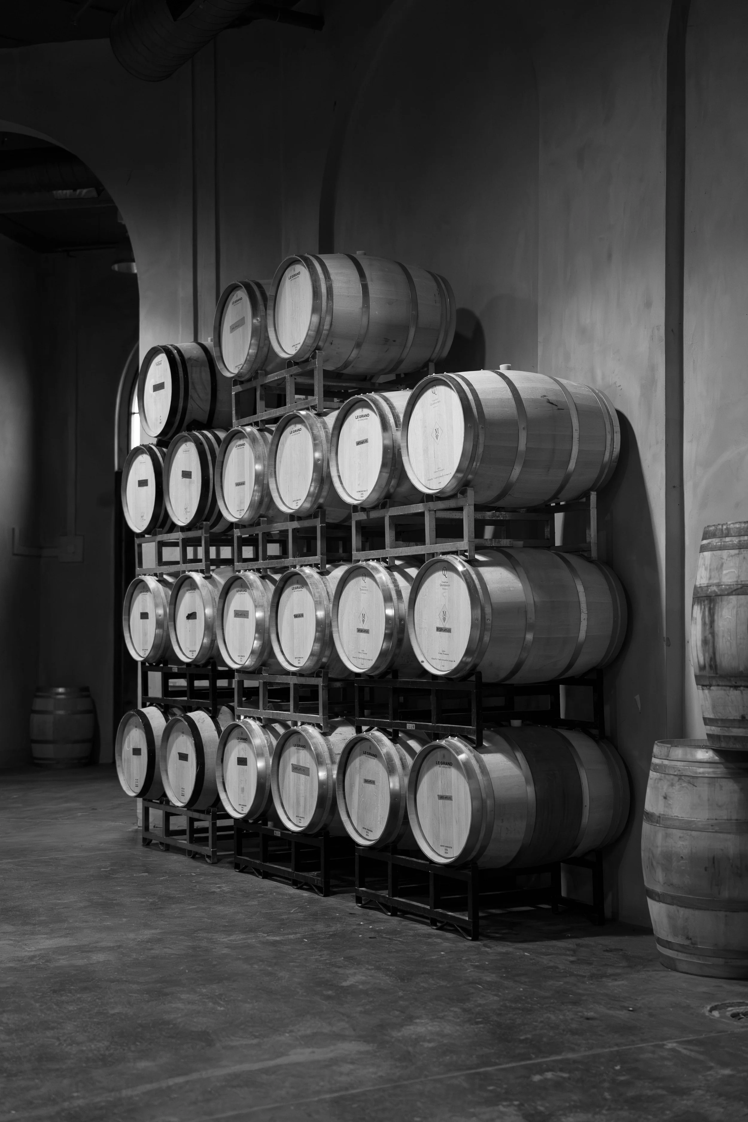 Black and white photo of wine barrels stacked on wooden racks in a winery cellar, with some barrels on the floor and arched windows in the background.