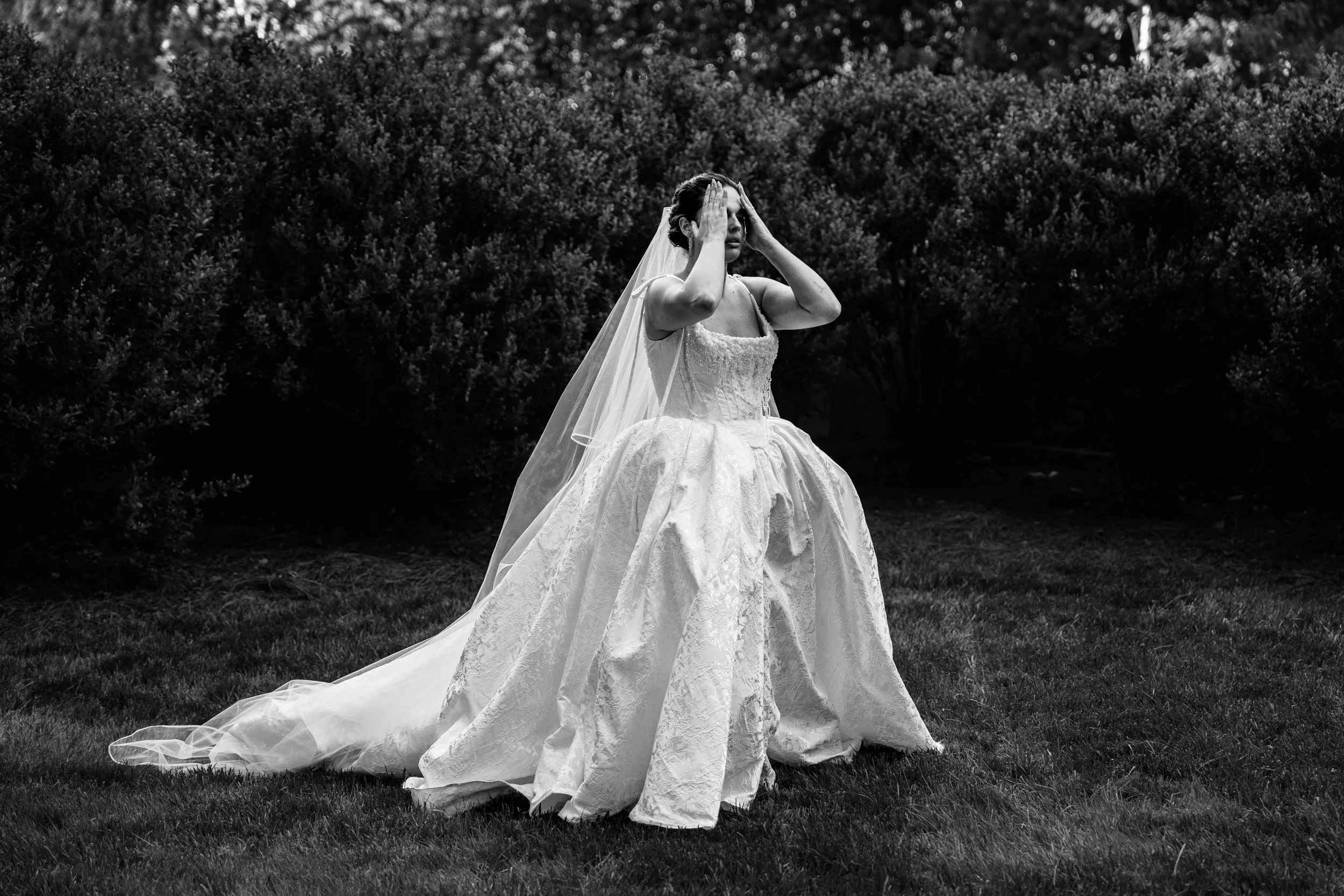 A woman in a wedding dress with a veil, standing on grass outdoors, holding her head with both hands, with dense bushes in the background, in black and white.