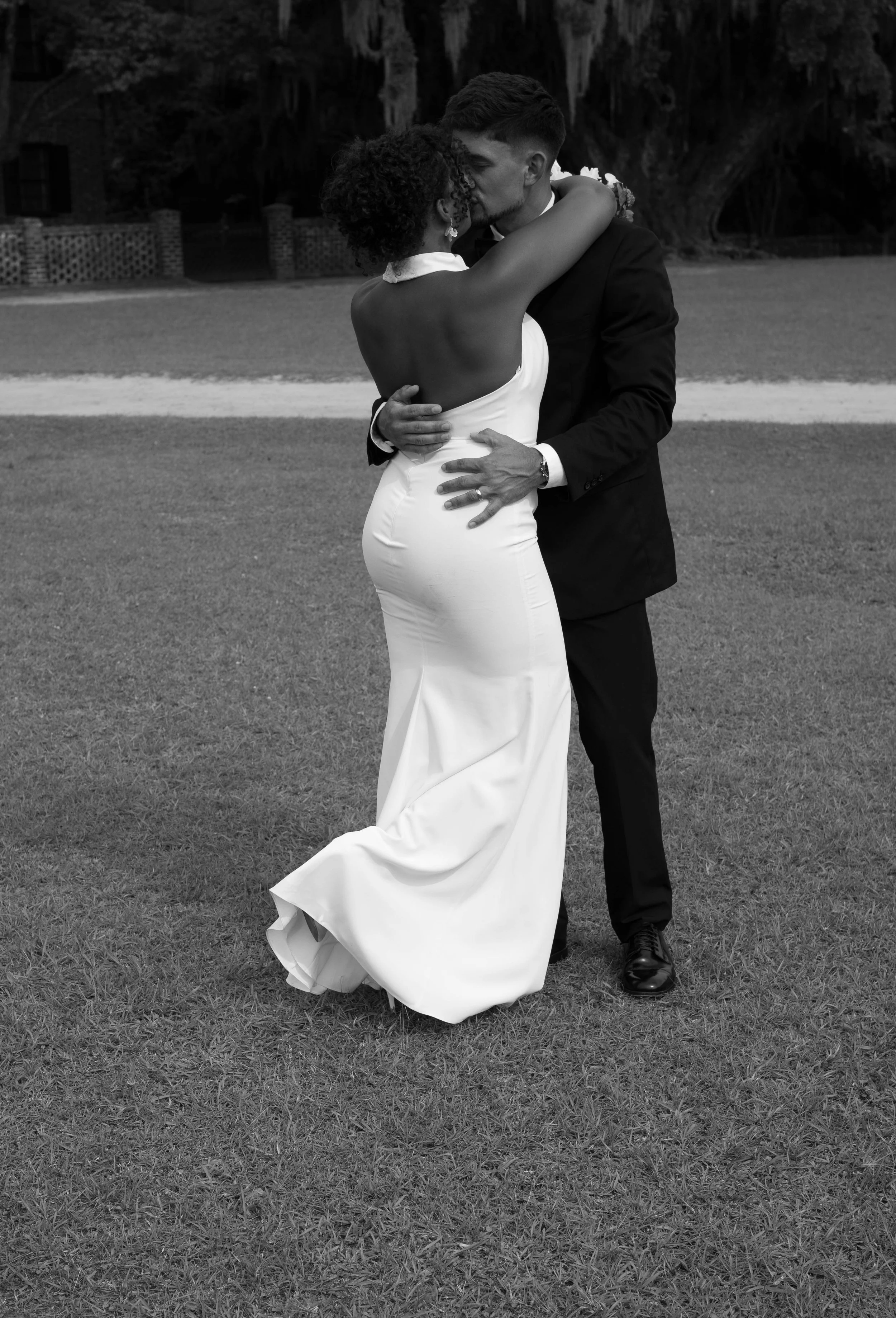 A couple embracing and sharing a kiss outdoors, woman in a long white dress and man in a dark suit, on a grassy lawn with trees in the background.