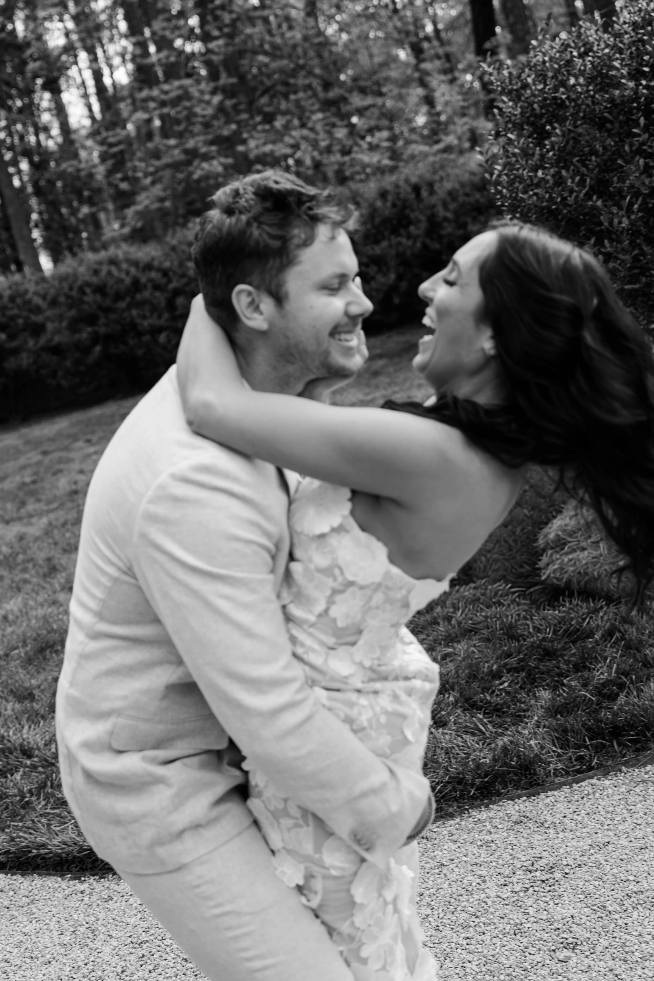 A black-and-white photo of a couple outdoors, smiling and looking at each other. The woman is wearing a dress with floral details, and the man is in a light-colored shirt. They are embracing with the woman's arm around the man's shoulder, surrounded by trees and bushes.