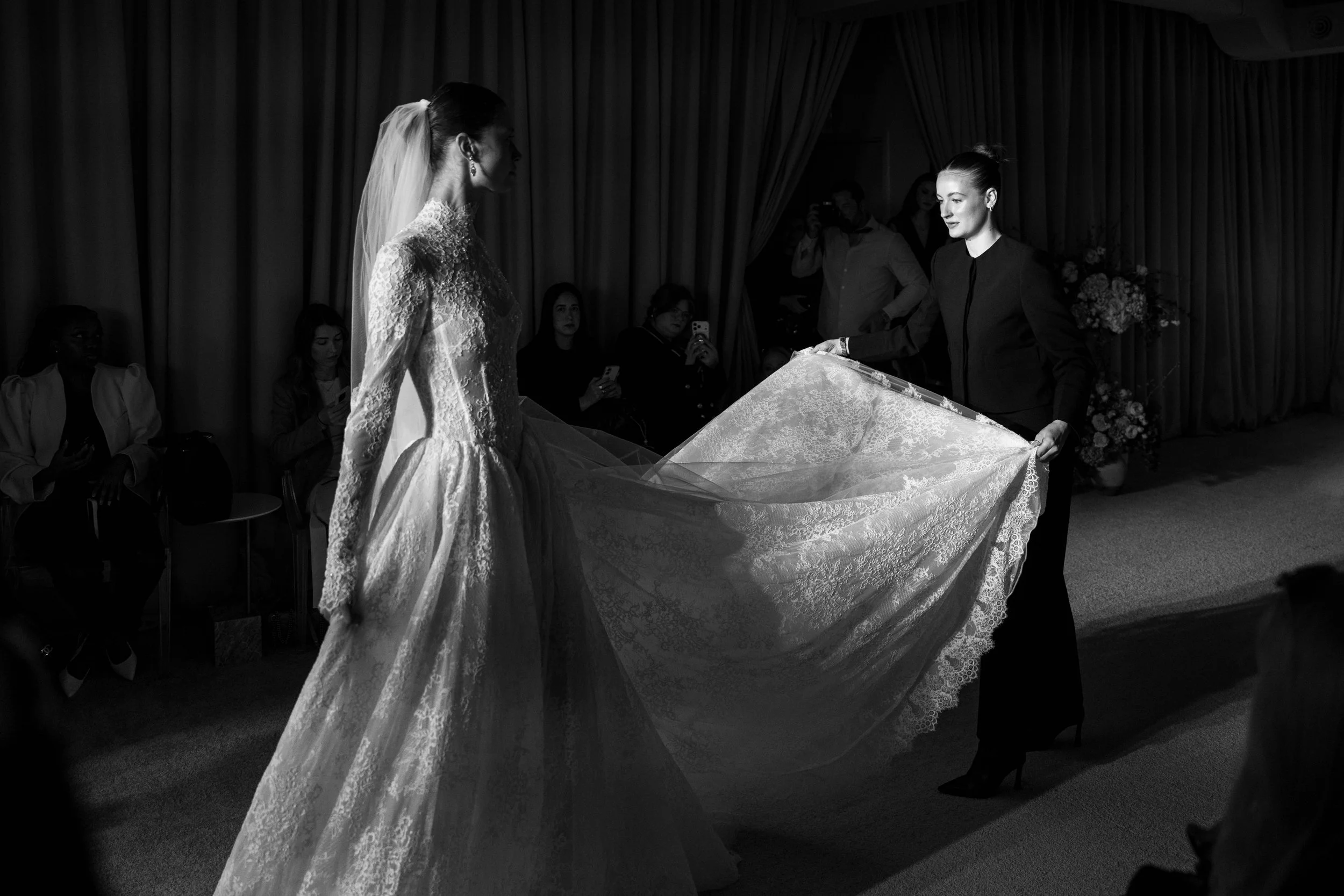 A black and white photo of a wedding ceremony, showing a bride in a lace wedding gown and a veil, facing a woman in a dark outfit, who appears to be reading or holding a cloth or lace. The background includes seated guests and curtains.
