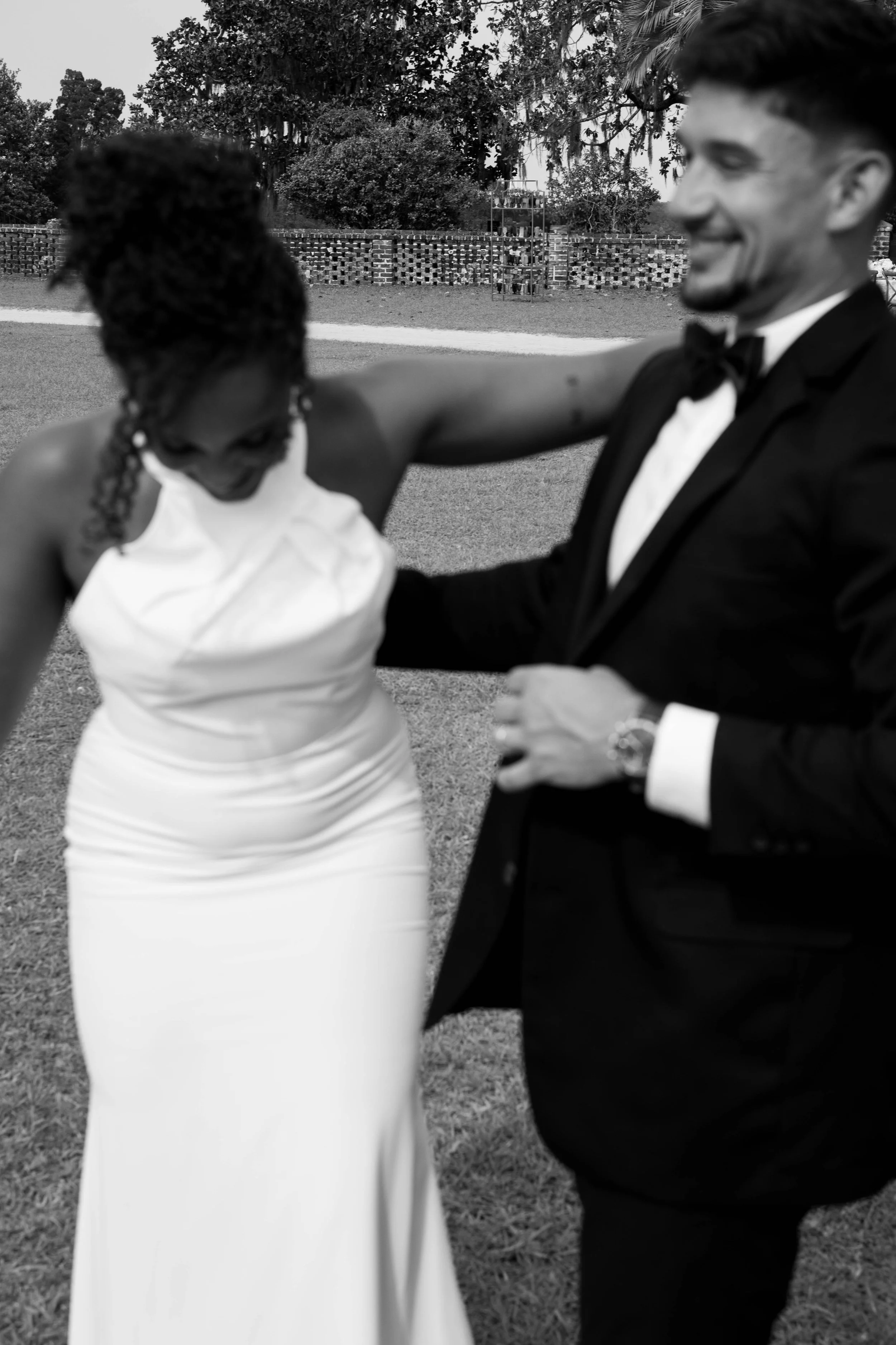 Black and white photo of a bride and groom celebrating outside, the bride in a white dress with her head bowed and the groom in a tuxedo smiling, as they dance together.
