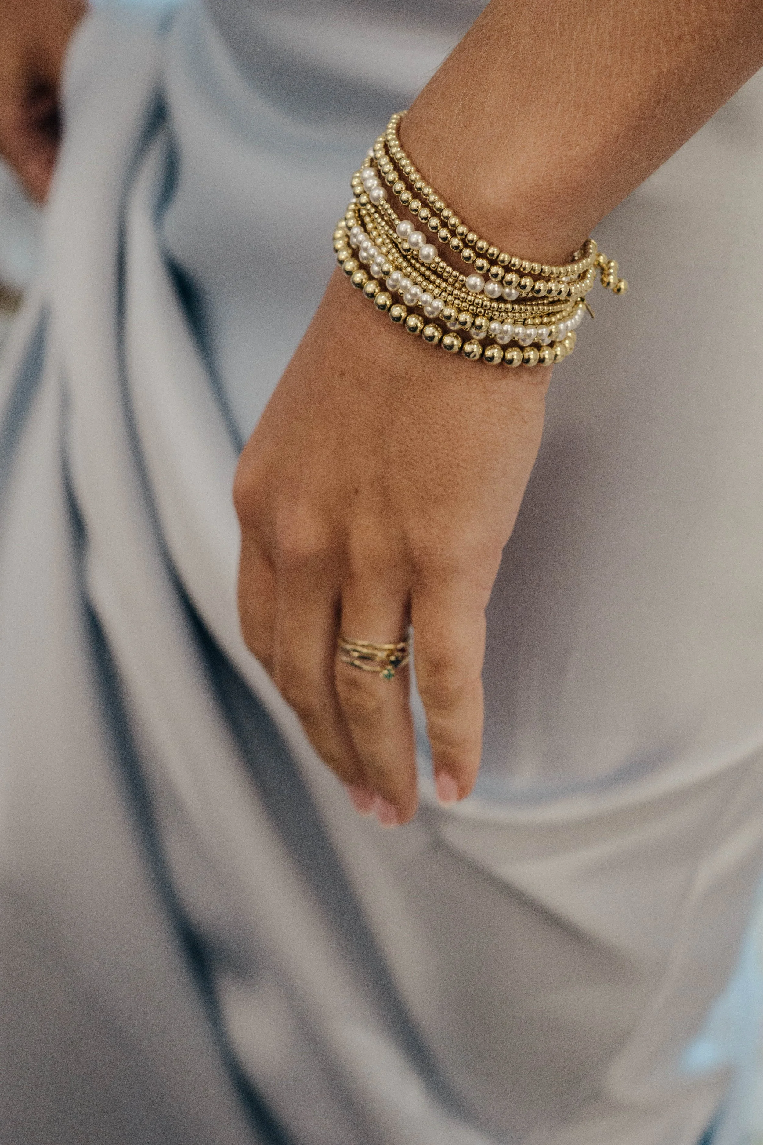 Close-up of a person's hand and wrist showcasing multiple gold and pearl bracelets, with a ring on their finger, against a background of light-colored clothing.