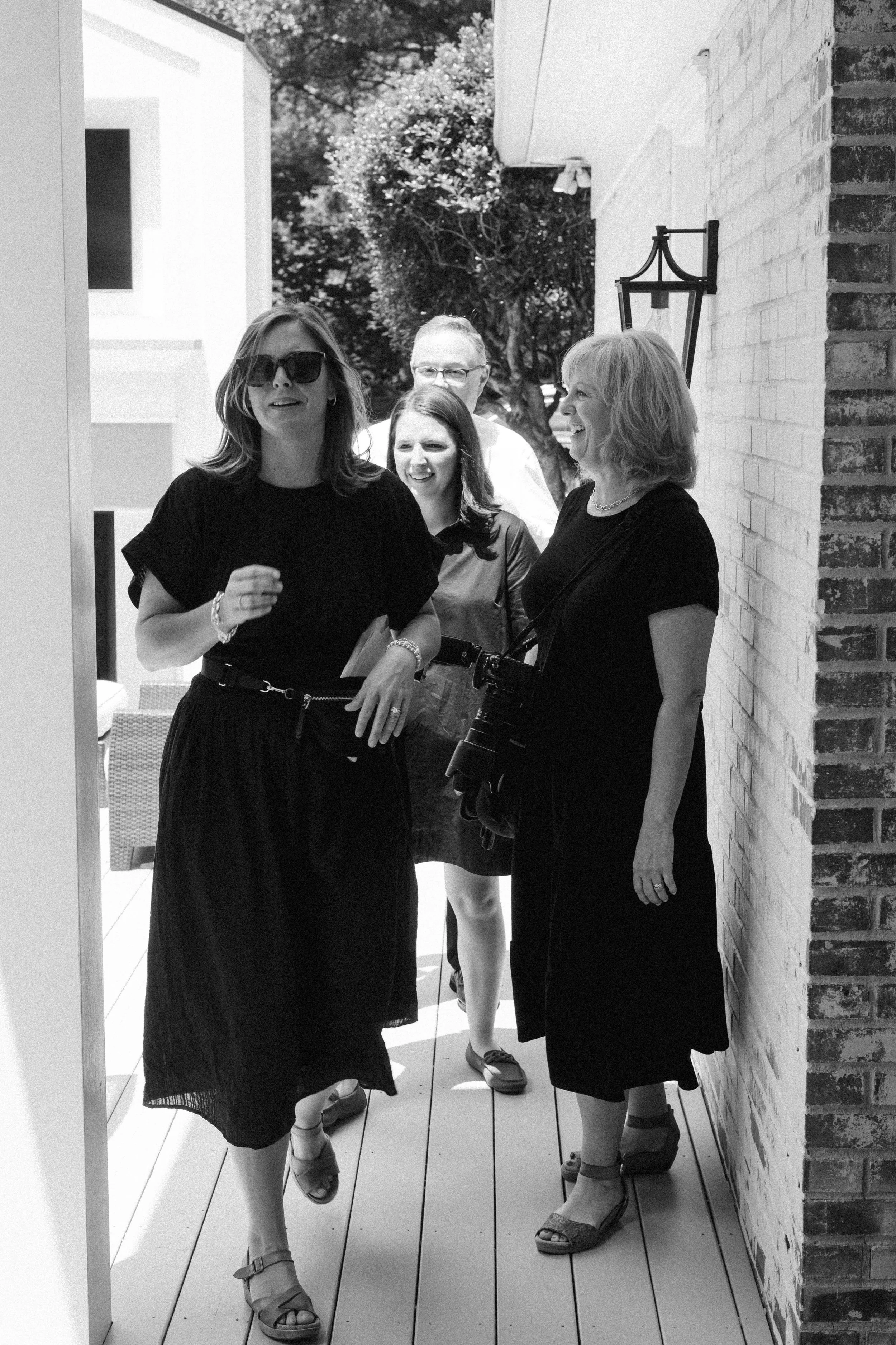 A group of four women walking outside on a sunny day, one woman wearing sunglasses and carrying a camera, smiling and talking.
