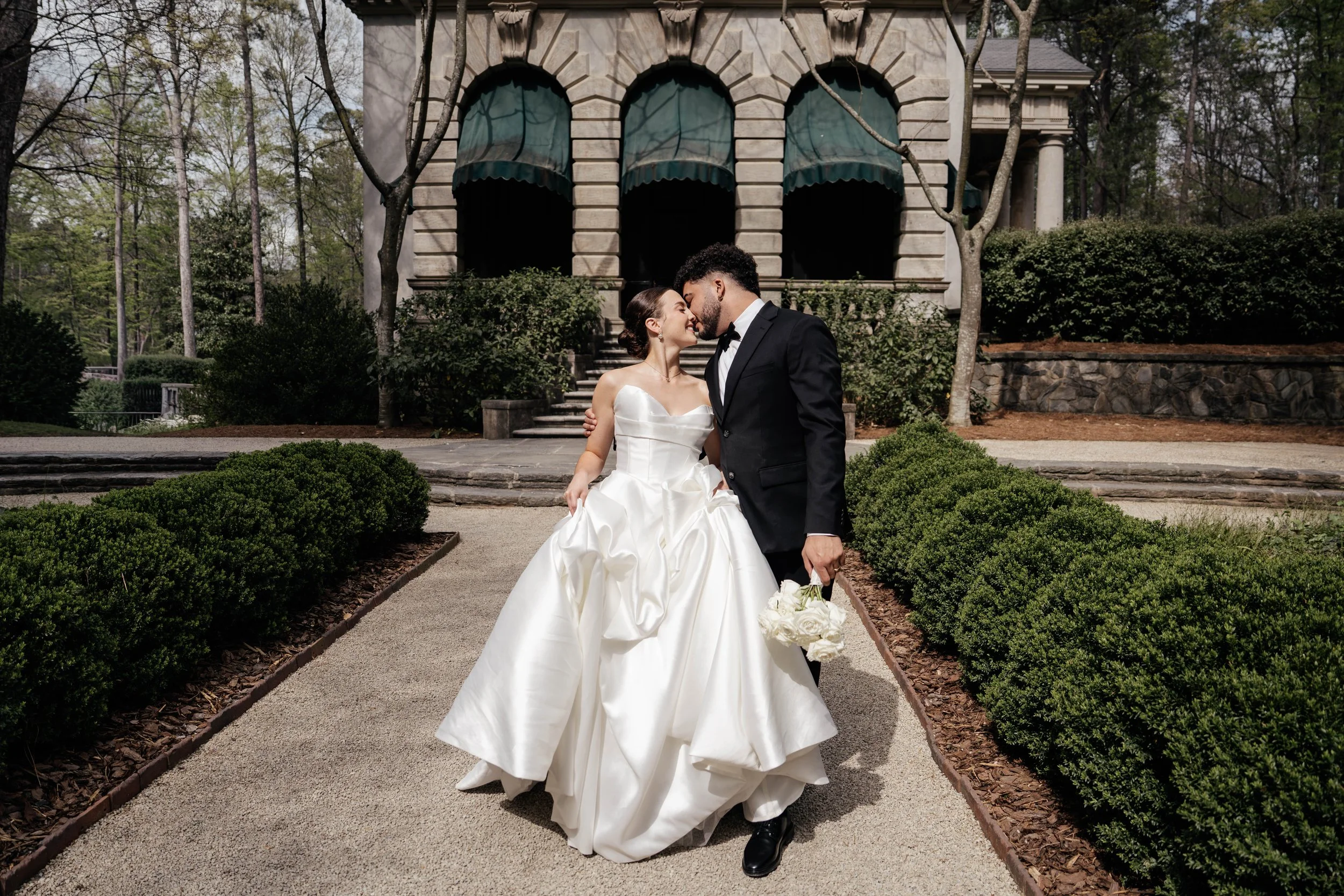 A bride and groom kissing in front of a historic building with arched windows, surrounded by trees and bushes.