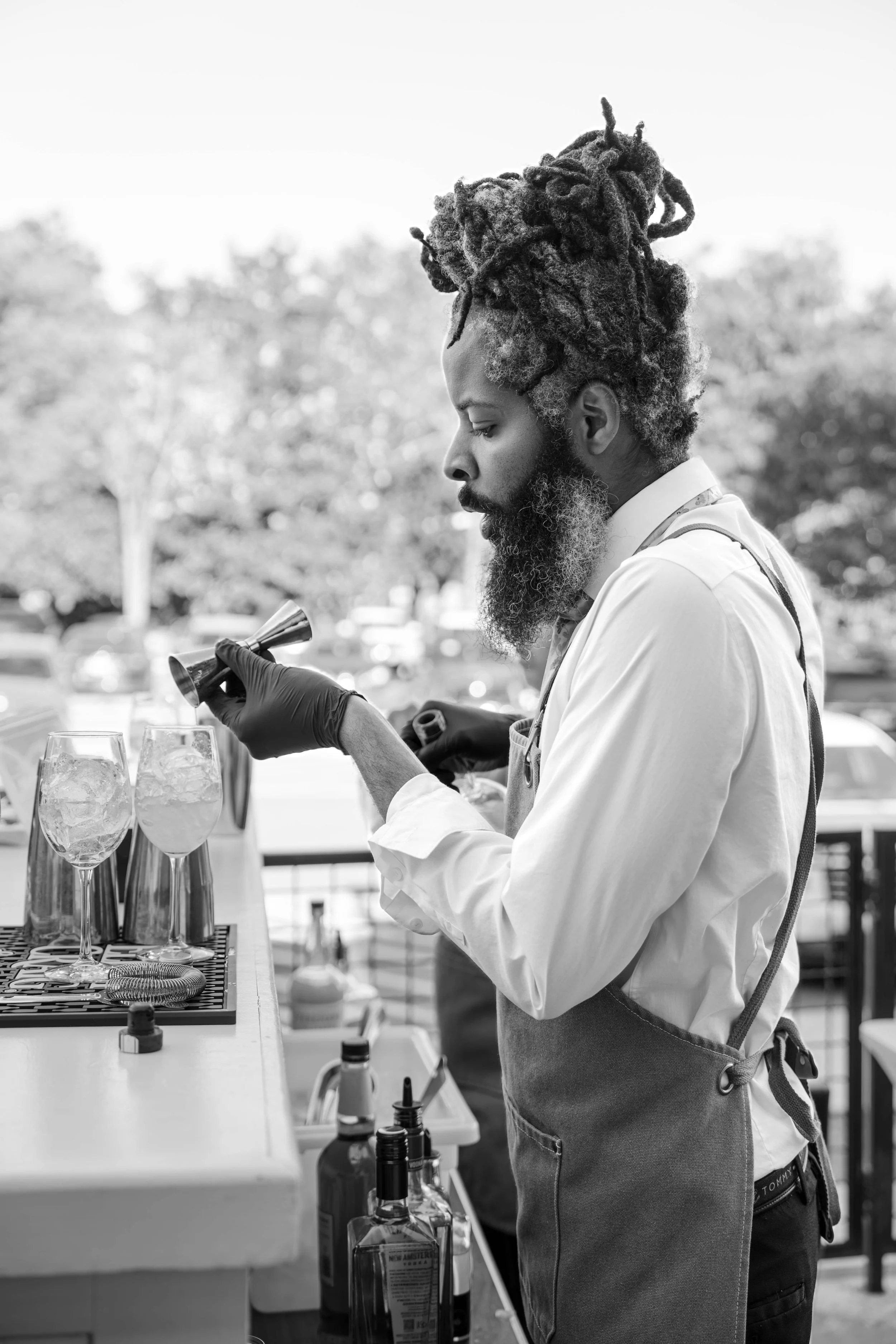 A man with a beard and dreadlocks, wearing a white shirt and an apron, is standing at a bar or outdoor table. He is holding a cocktail shaker with gloves on, preparing drinks. Several glasses with ice and bottles are on the table. The background show