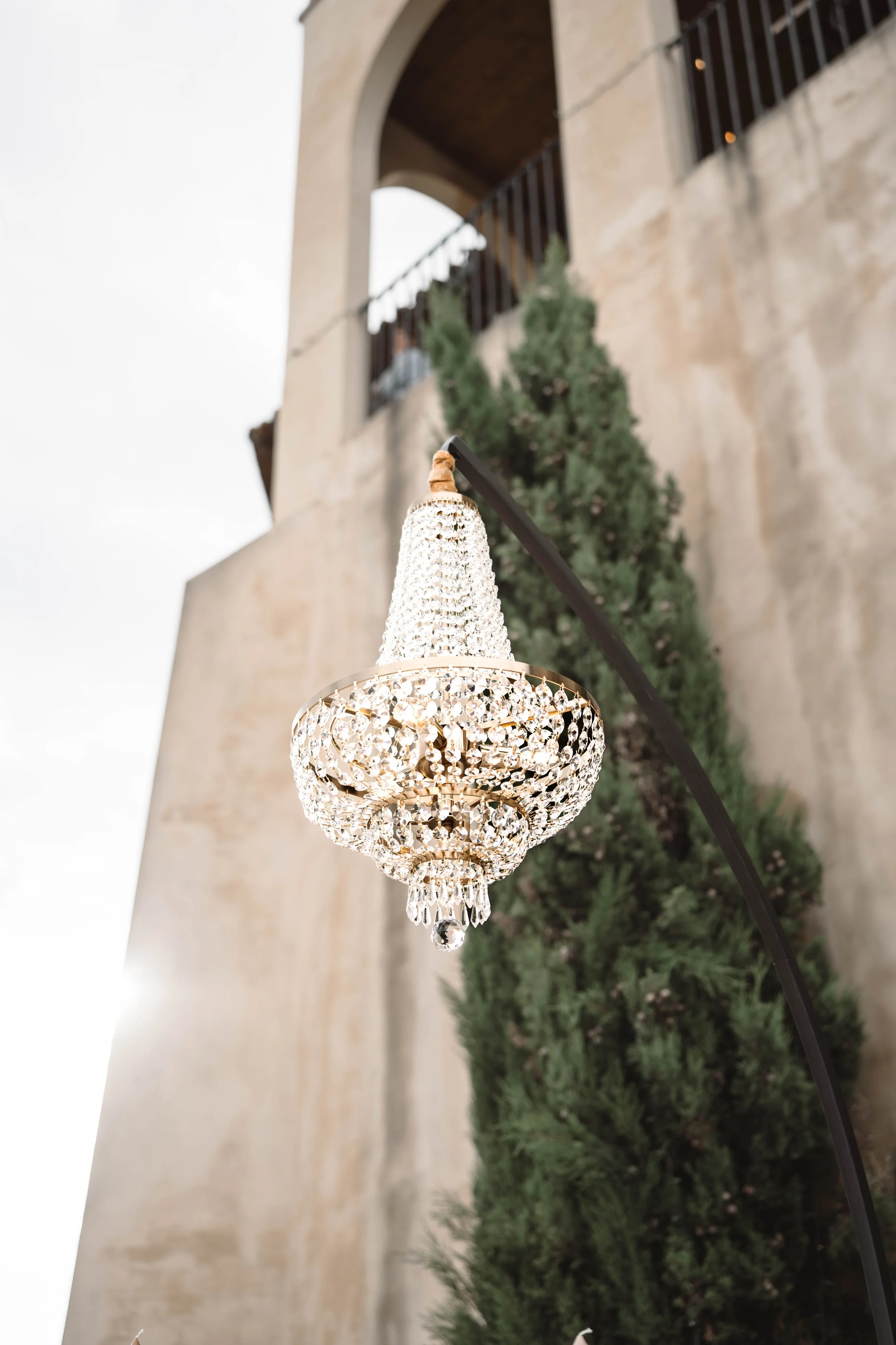 A luxurious chandelier hanging outdoors near a stucco wall and a green shrub, with large arches and a balcony in the background.