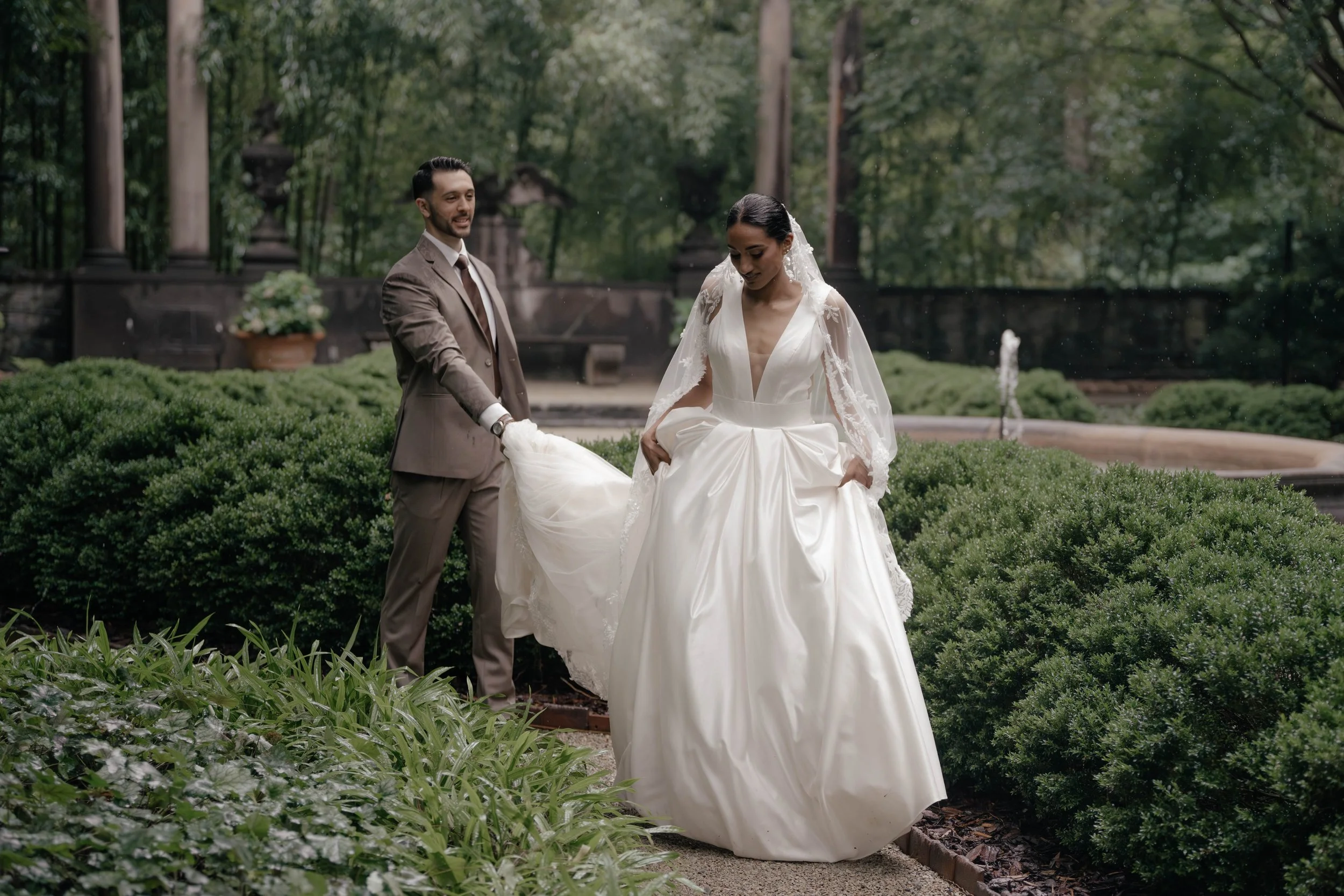 A bride and groom standing outdoors in a lush, green garden. The groom is wearing a brown suit and holding the bride's dress, while the bride is dressed in a white wedding gown with lace details and a veil, looking down as she walks.