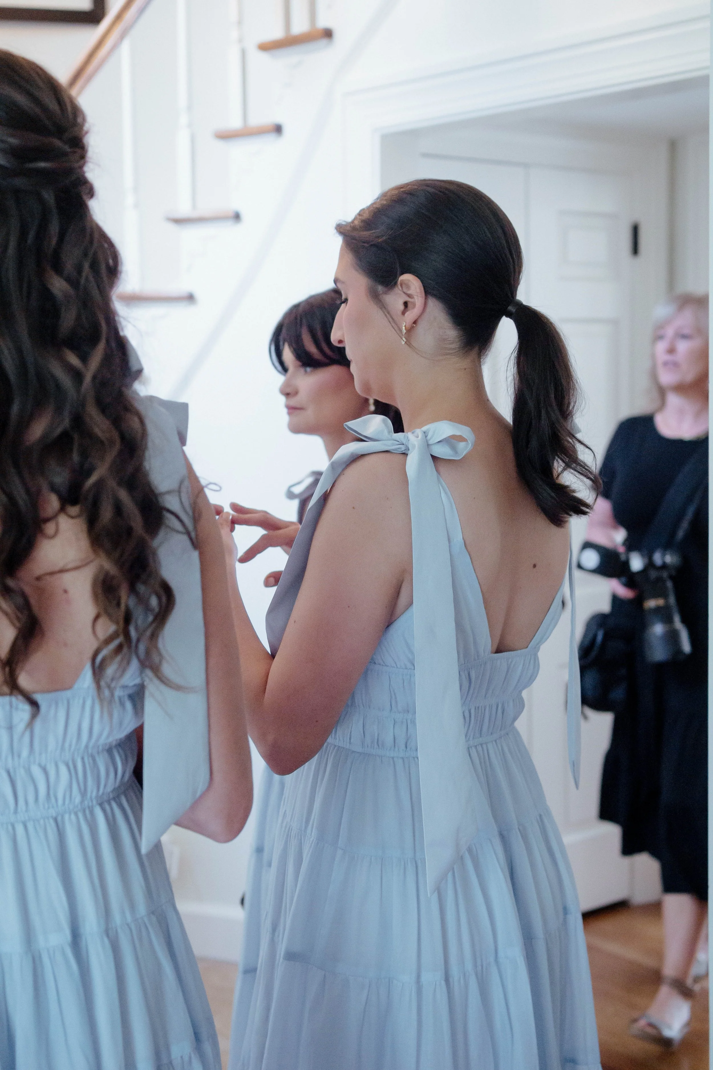 Women in matching light blue dresses preparing for an event indoors, with a woman in black attire holding a camera in the background.