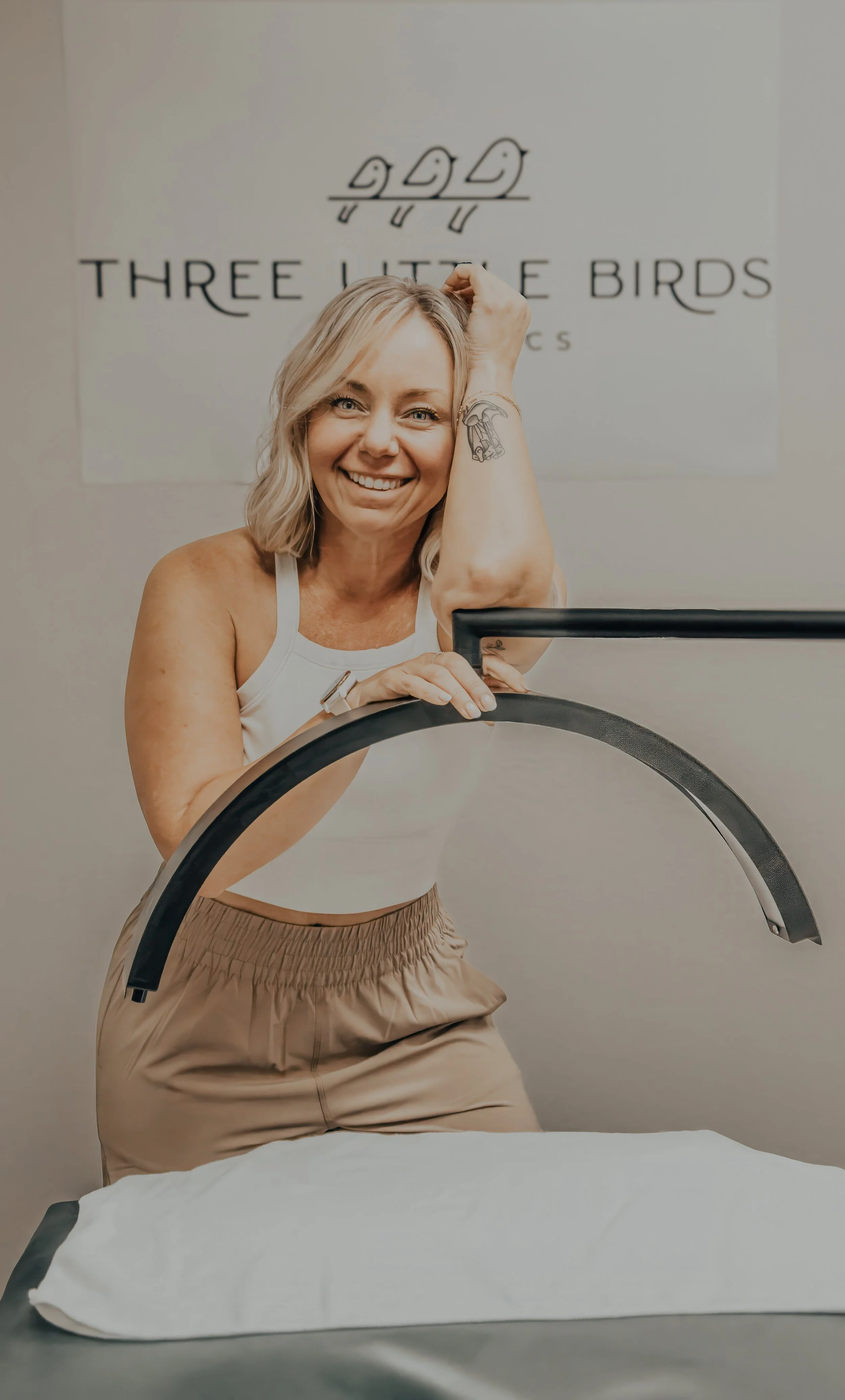 A smiling woman in workout attire leaning on a table with a black object in her hand, in front of a sign that reads 'Three Little Birds' with a bird illustration.