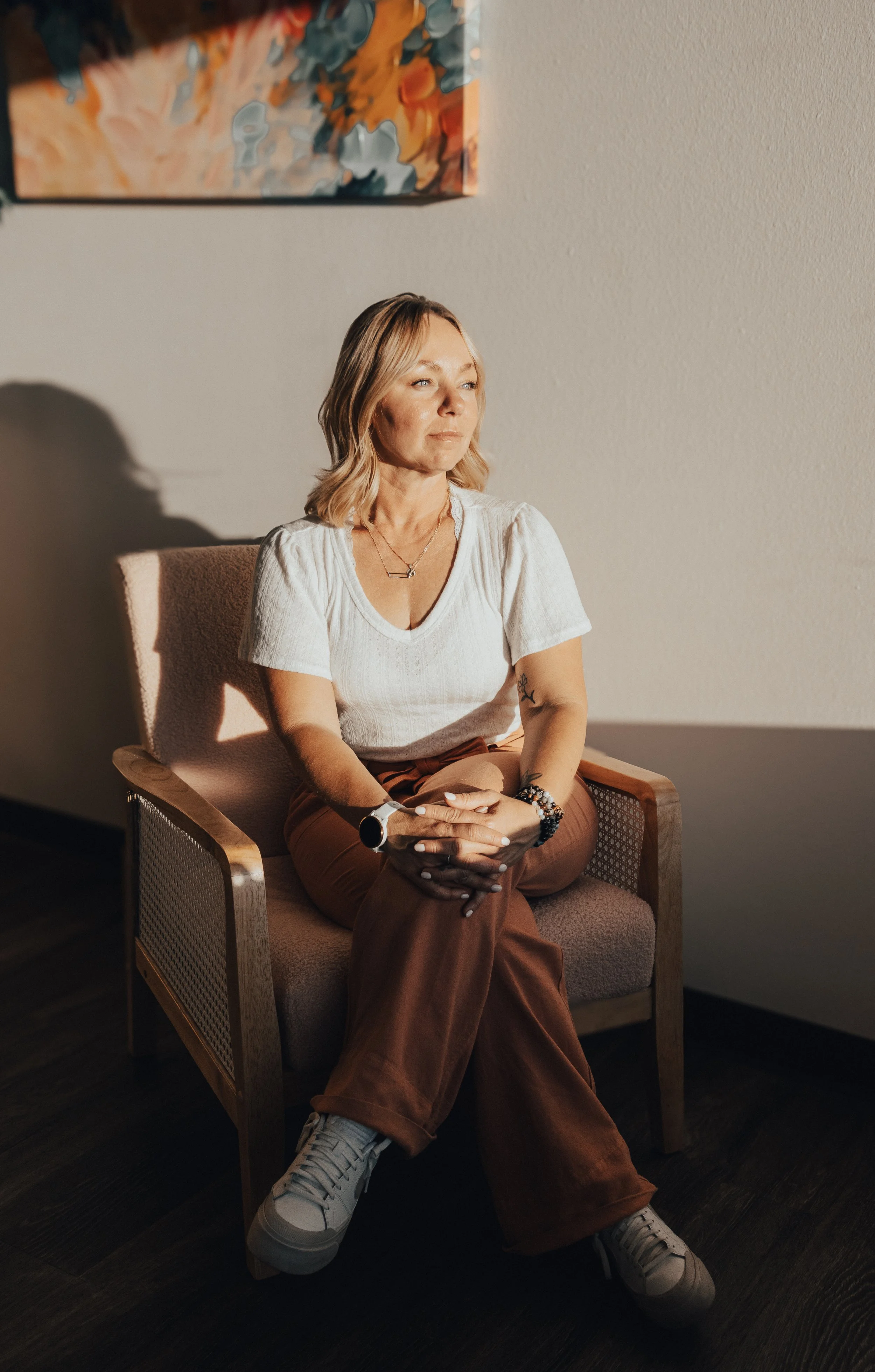A woman with blonde hair, wearing a white t-shirt, brown pants, and sneakers, sitting on a beige chair with wooden armrests in a room with a plain wall and an abstract painting. She is looking to her right with her hands clasped in her lap.