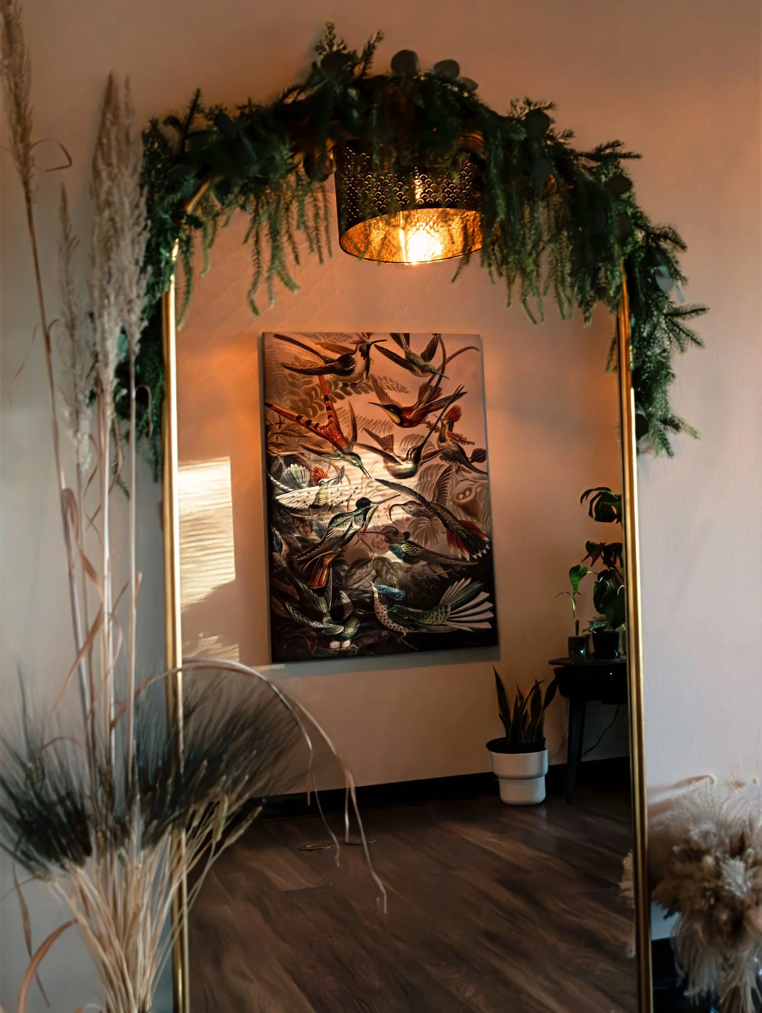 Decorative mirror with a green garland border reflecting a room with a hanging light, a framed bird artwork, and potted plants.