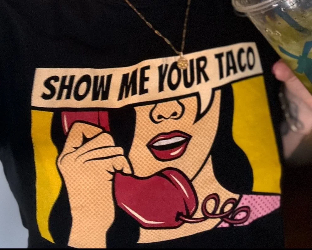 A person wearing a black shirt with comic-style art of a woman holding a red phone and speaking into it, with a speech bubble saying 'SHOW ME YOUR TACO'.