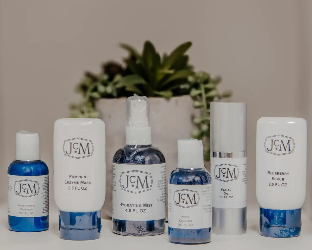 Skincare product bottles including moisturizer cleanser, pumpkin enzyme mask, hydrating mist, toner, facial oil, and blueberry scrub, arranged in front of a potted succulent plant.