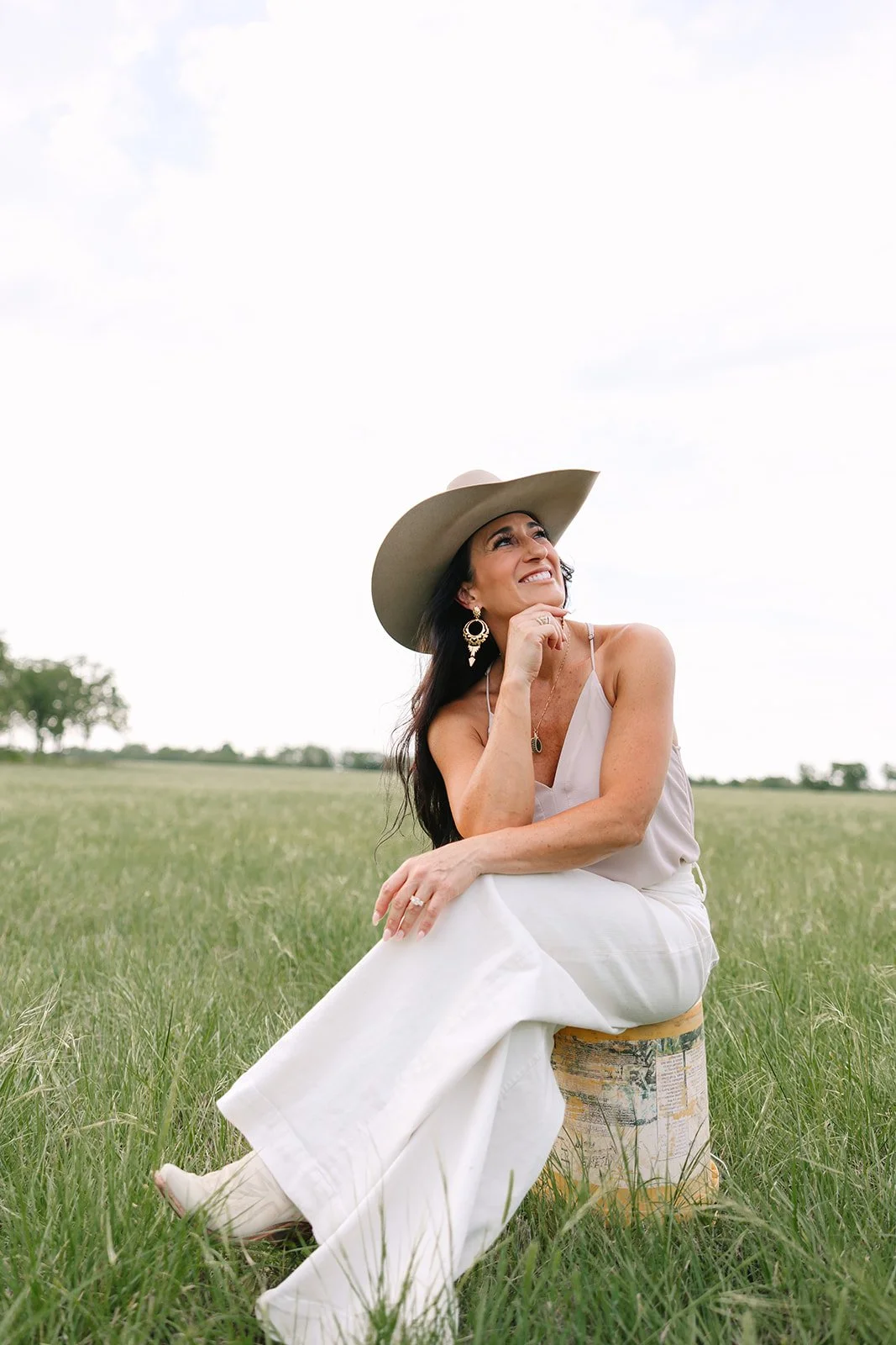 Woman in wide-brim hat and white outfit sits on a stool in a grassy field, smiling thoughtfully and looking upward.