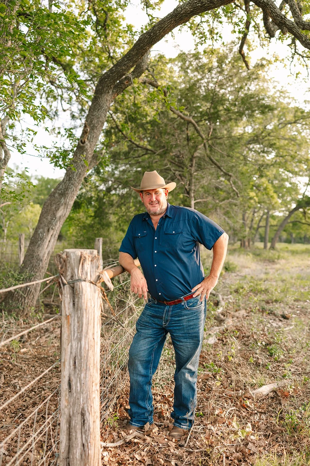 Man in a cowboy hat leans on a wooden fence post in a rural outdoor setting with trees and grass around him.