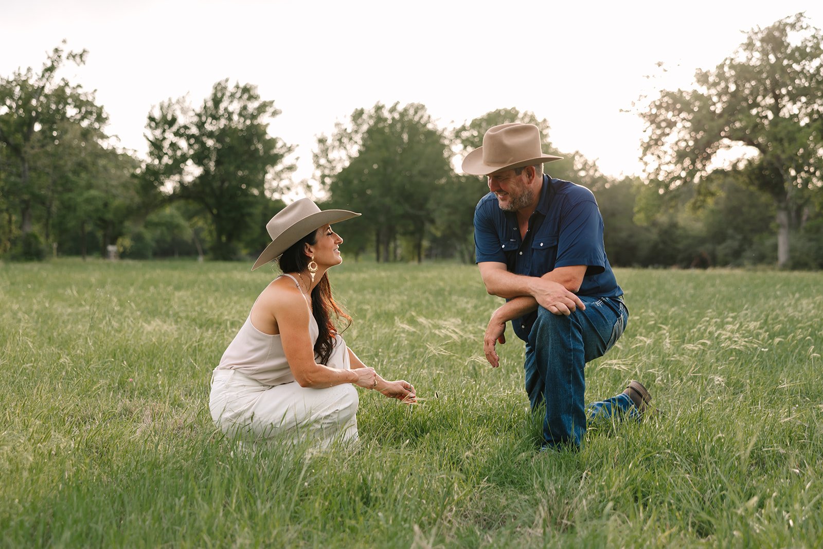 A woman and a man in cowboy hats talk together in a grassy field, surrounded by trees on a sunny day.