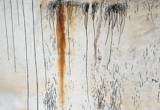 A close-up of a weathered surface with vertical black and rust-colored streaks, creating an abstract and distressed texture against a beige background, evoking an industrial or aged aesthetic.