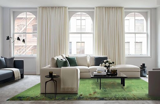 A bright, modern living room with large arched windows, cream curtains, and a cozy white sectional adorned with green and gray pillows. A green rug adds a pop of color beneath a sleek black coffee table. A minimalist side table holds a candle.