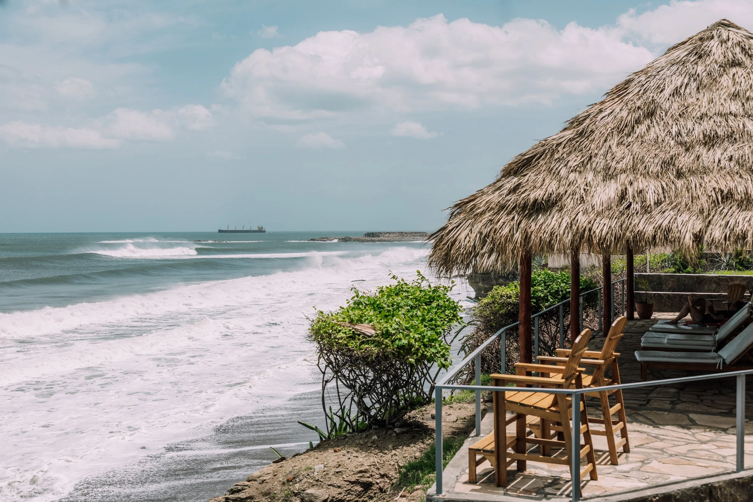 Oceanside view with waves crashing, a thatched-roof hut with patio chairs, and a cargo ship in the distance under a partly cloudy sky.