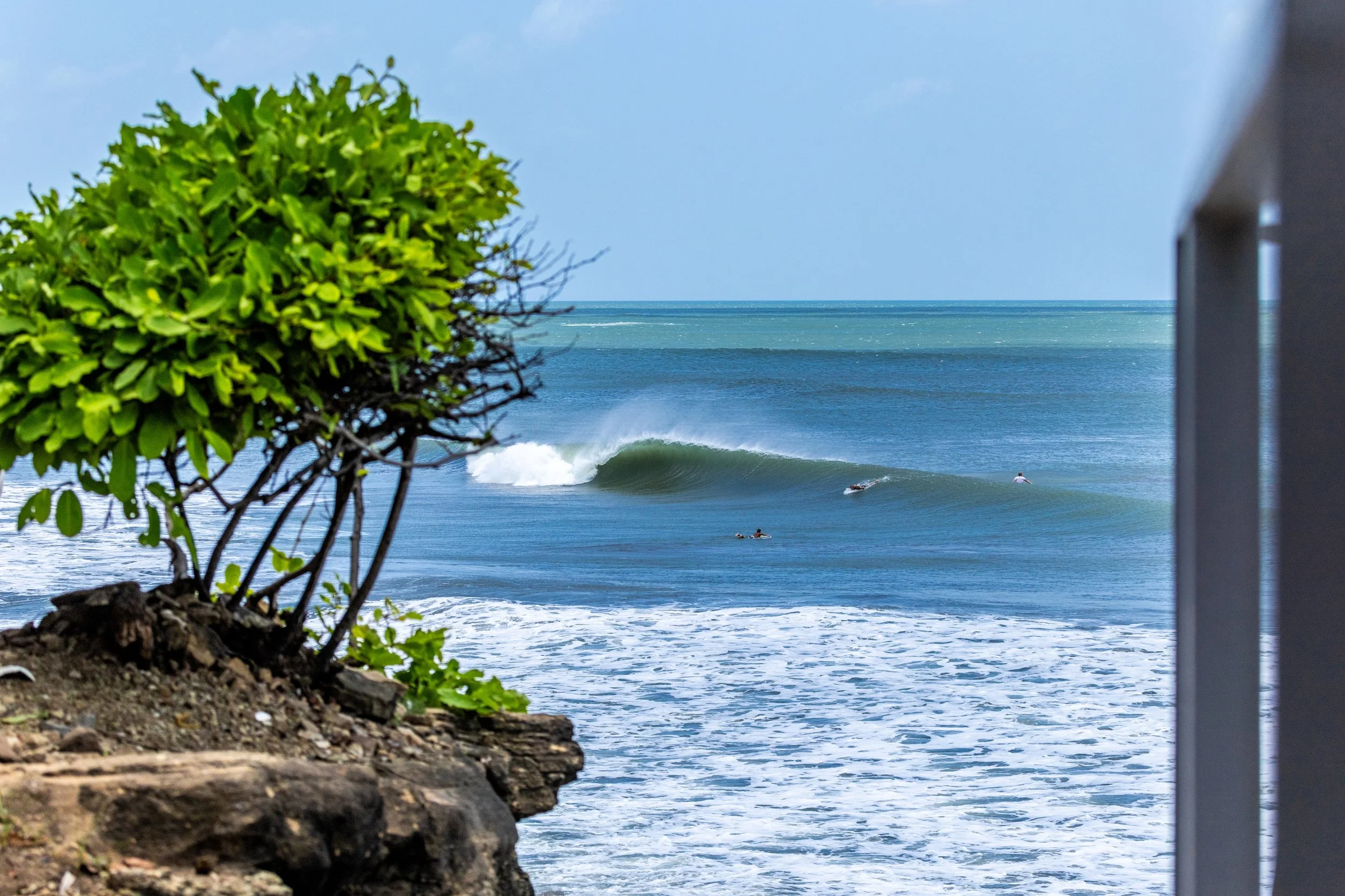 People surfing and swimming in ocean waves at the beach during daytime, with a large wave and clear blue sky in the background.