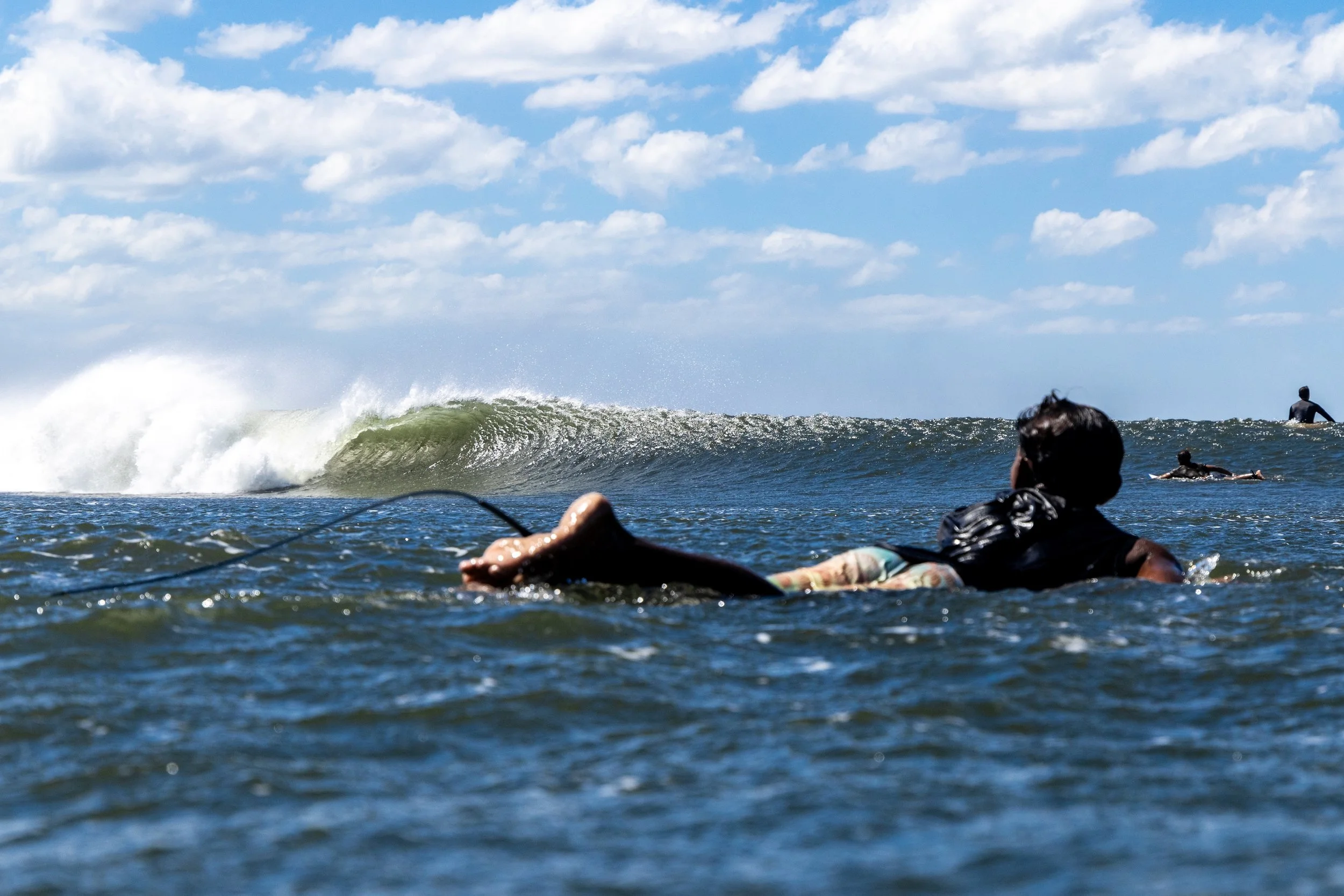 People surfing and swimming in the ocean under a blue sky with clouds.