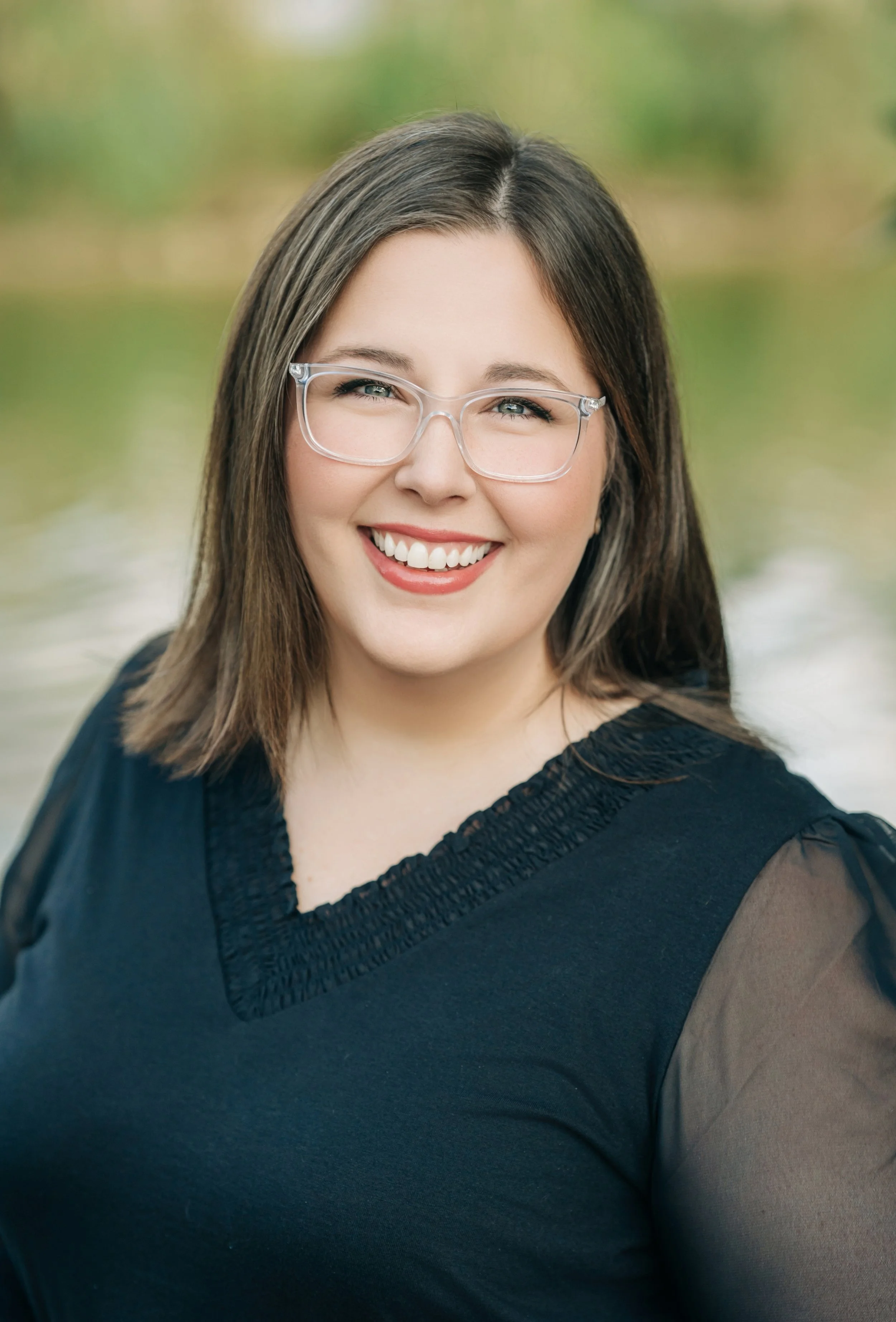 A smiling woman with shoulder-length brown hair, wearing glasses and a black top, outdoors with blurred green and brown background.