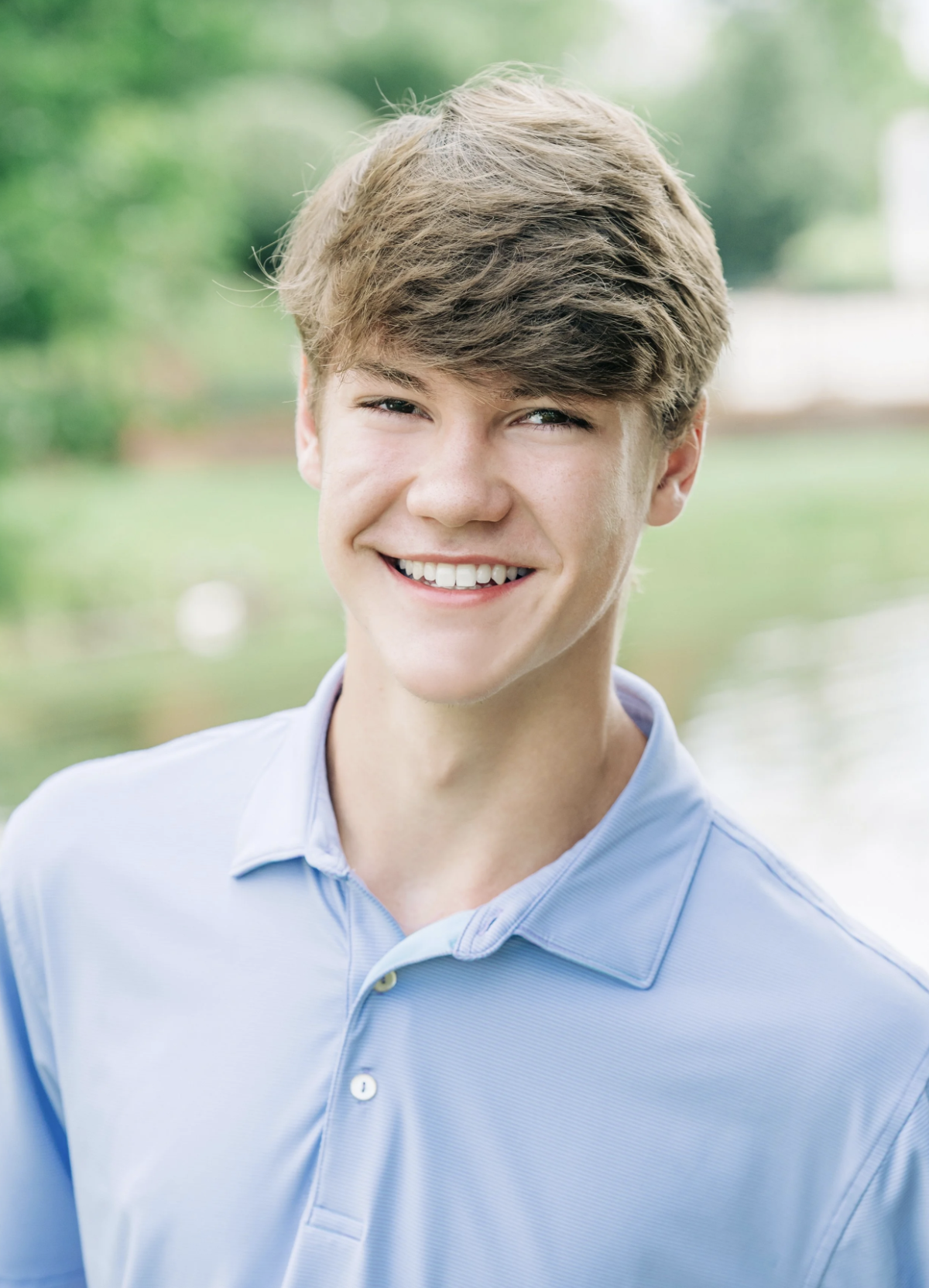 A young man with light brown hair smiling outdoors in front of a blurred green background, wearing a light blue button-up shirt.