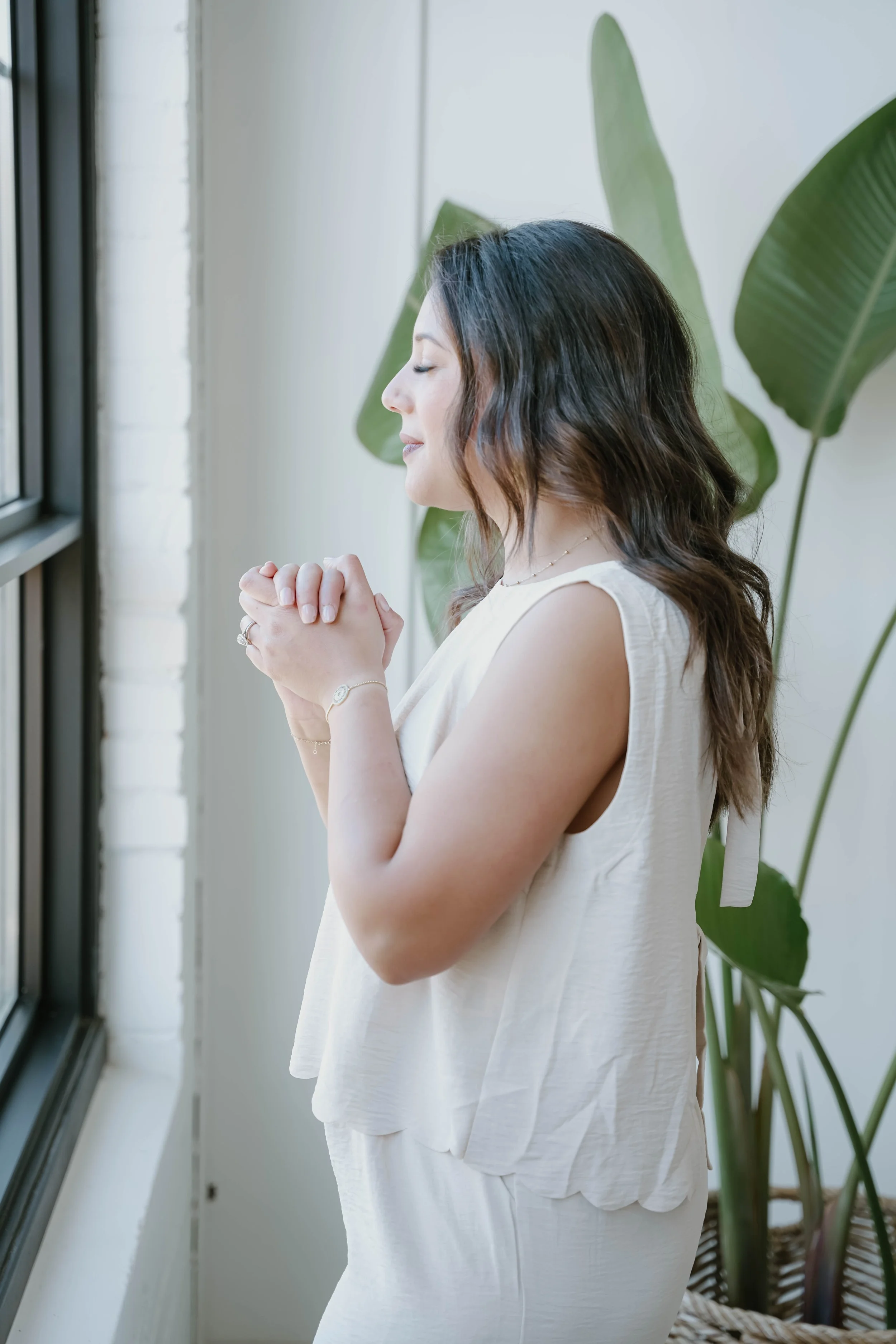 A woman with dark wavy hair and light skin standing with her eyes closed and hands clasped near her chest, facing a window with large green leafy plants in the background.