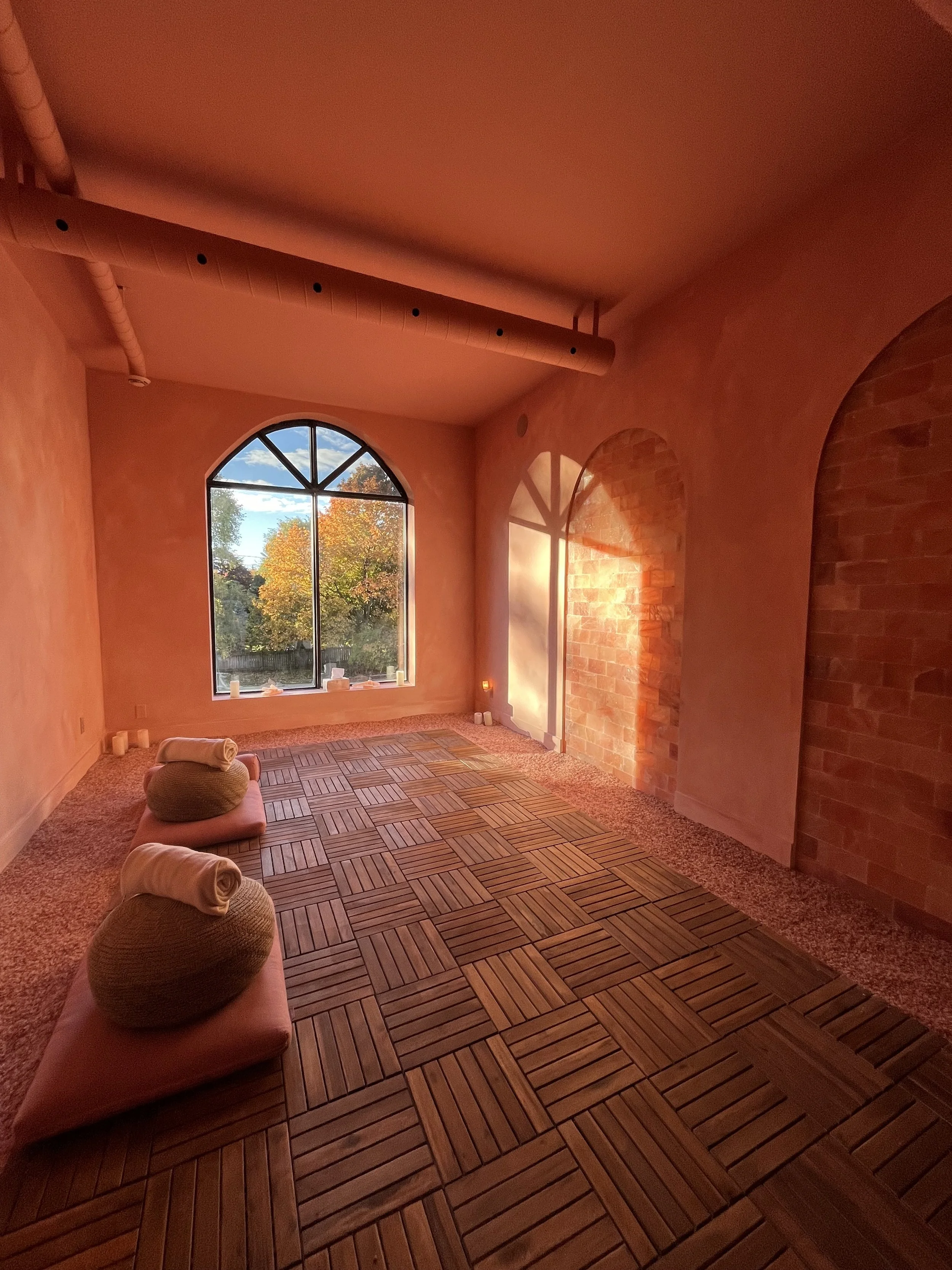 Sunlit room with wooden floor, pink walls, large arched window showing trees with fall foliage, and a cozy corner with rolled towels on cushions.