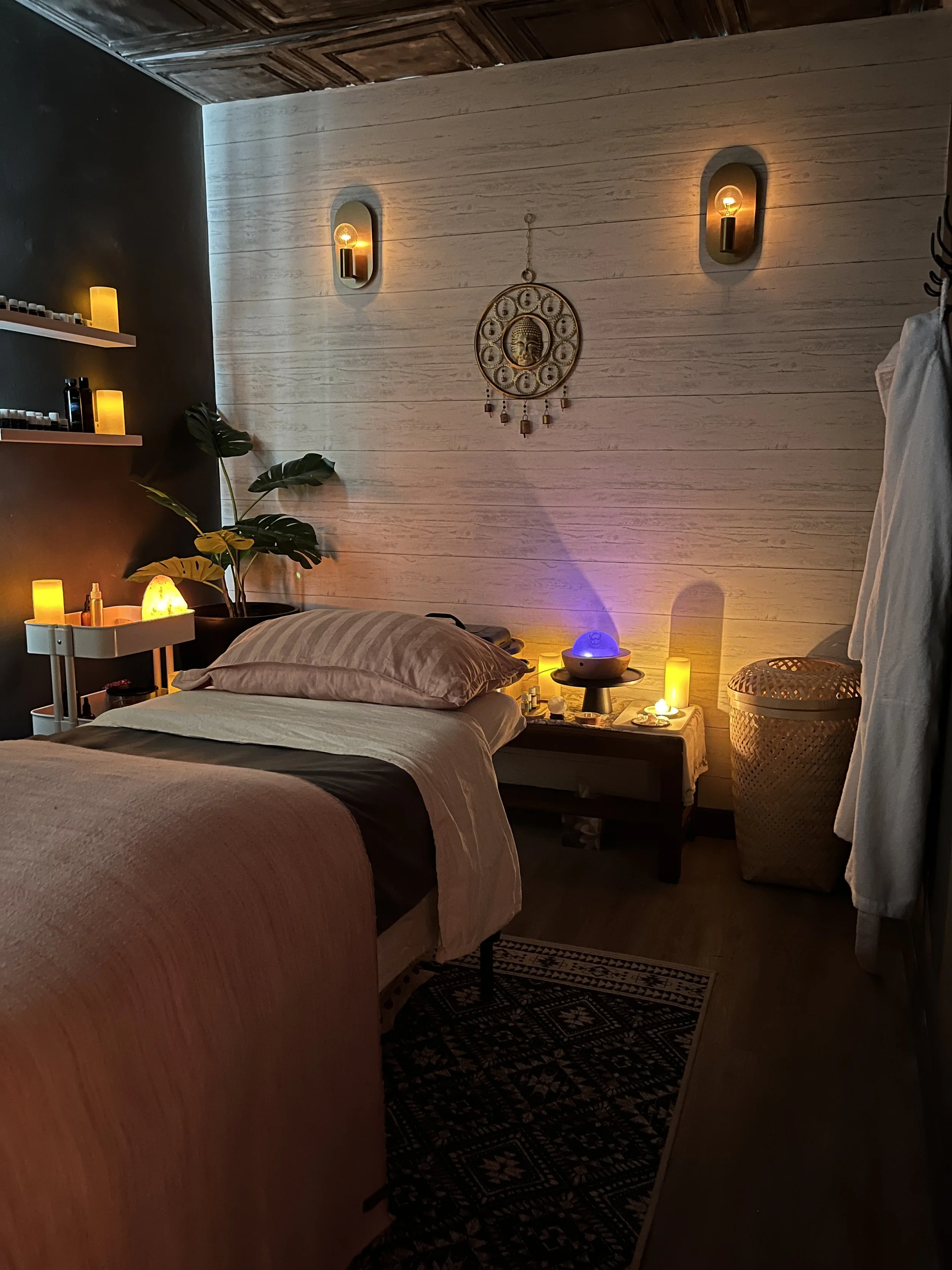 A cozy bedroom with warm lighting, a large bed with plush pillows, a black nightstand with a salt lamp and other items, a leafy plant, decorative candles, a large laundry basket, and a white wall with wall-mounted light fixtures and a bohemian-style wall hanging.