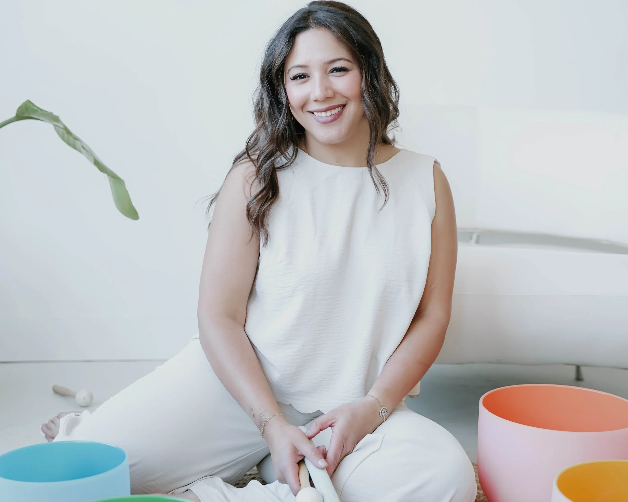 A woman with wavy dark hair smiling, sitting on the floor with colorful singing bowls around her, in a bright room.