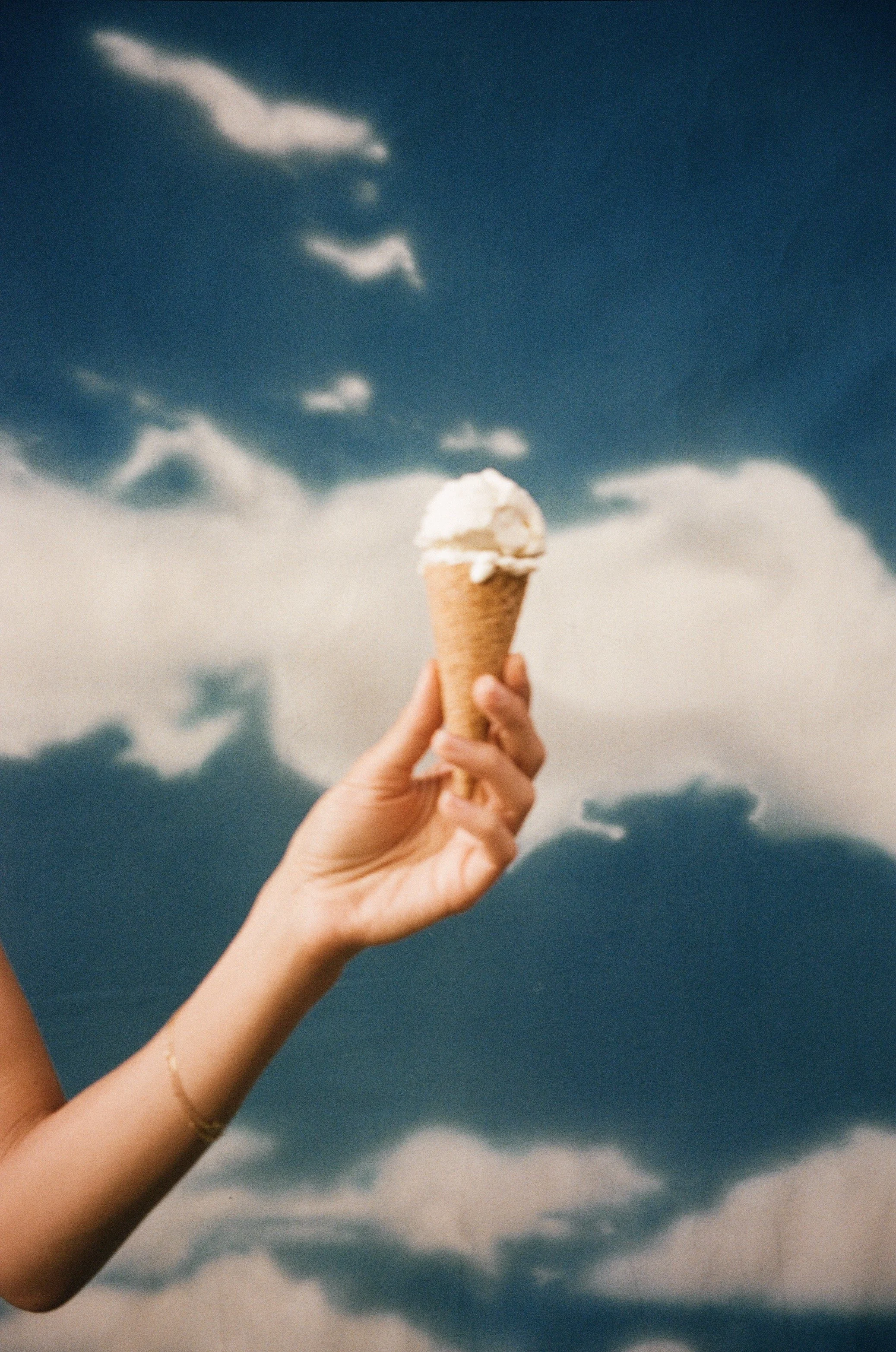 A person's hand holding an ice cream cone with a cloudy sky in the background.