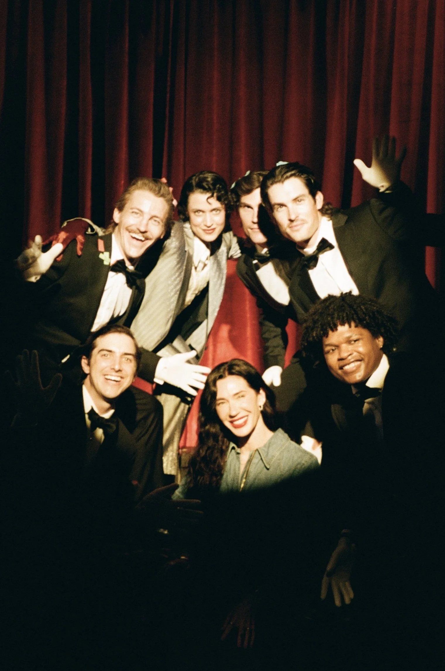 A group of people dressed in formal tuxedos and gowns, posing together on stage with red curtains in the background. They are smiling and making playful gestures.