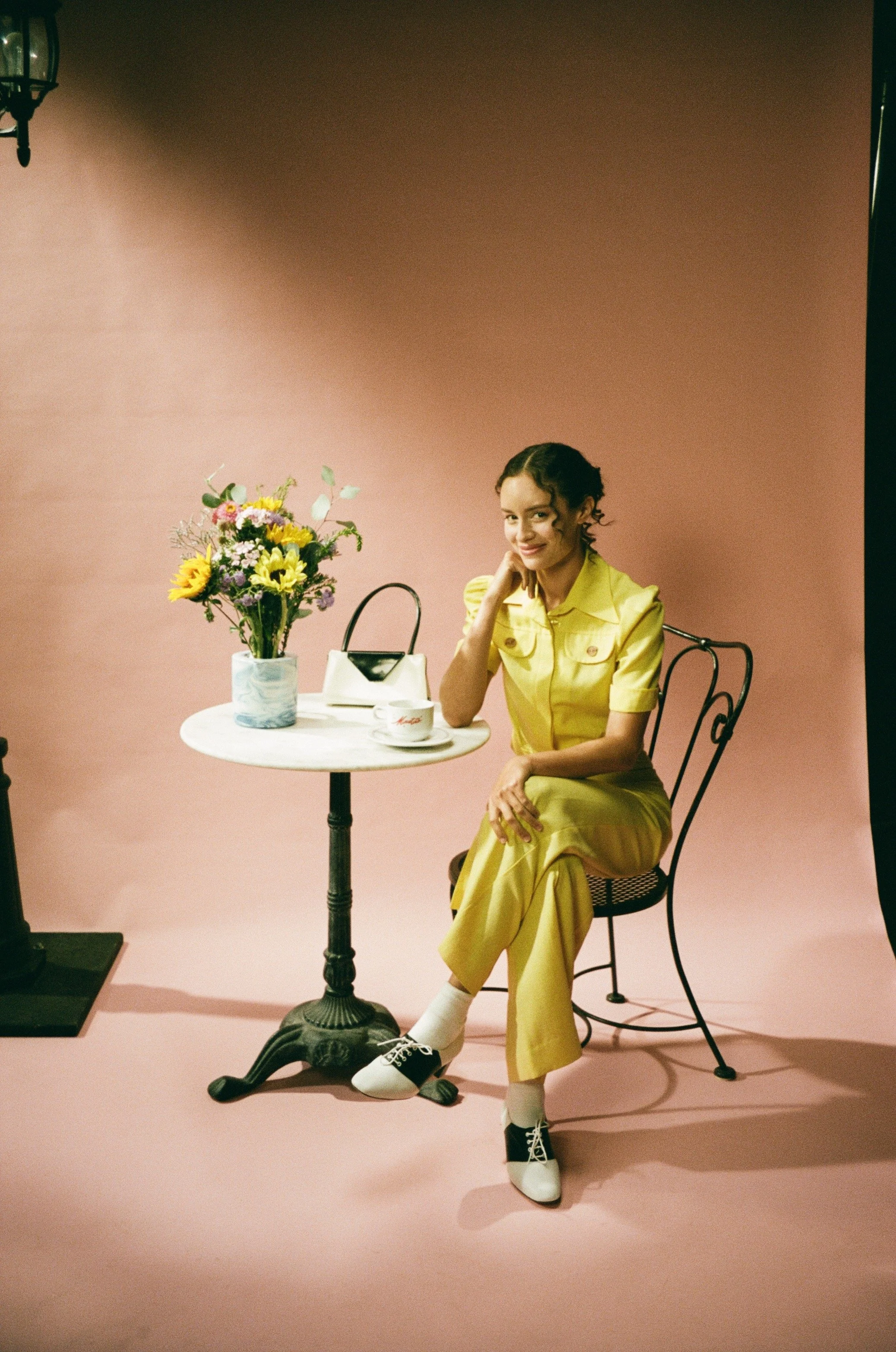 Young woman with curly hair in a yellow outfit sitting at a pink themed photo studio set with a pink background, a small round table with a flower vase, a purse, a mug, and a cup, sitting on a decorative black chair, smiling at the camera.