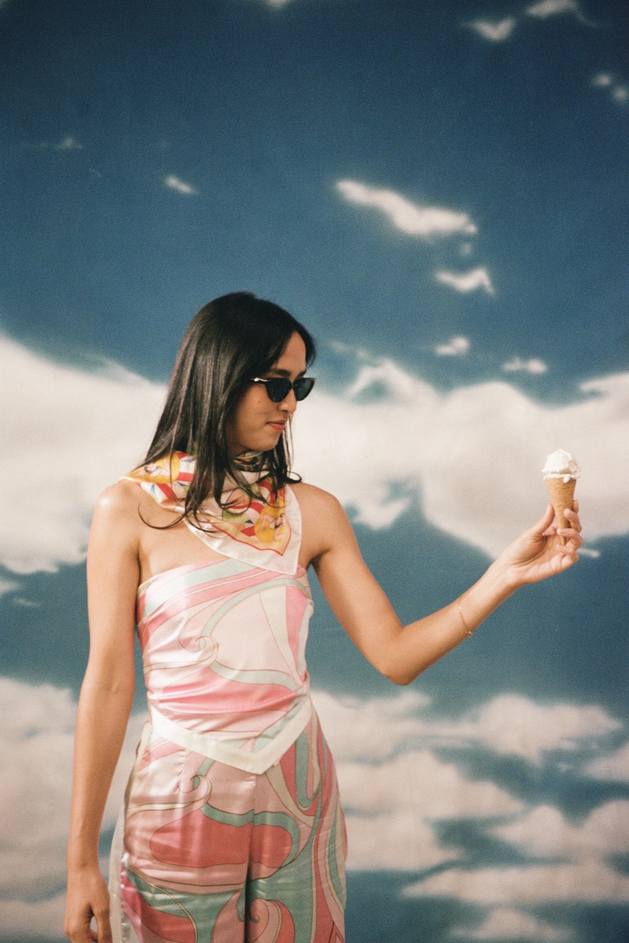 A woman with long dark hair wearing sunglasses and a patterned dress is holding an ice cream cone against a background of a cloudy sky.