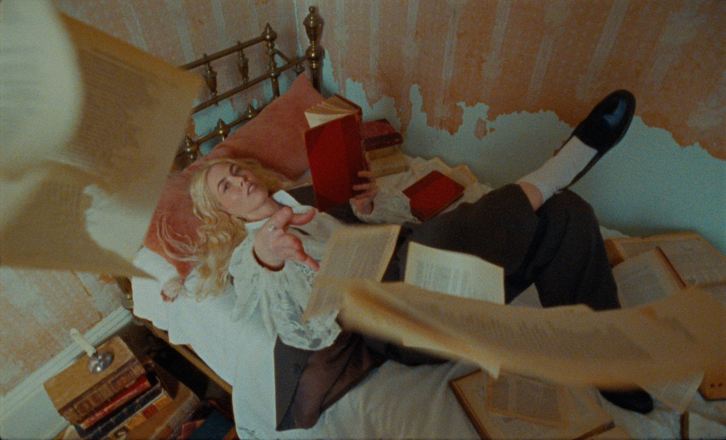 A woman with blonde hair is sitting at a wooden table, holding a few books. She is wearing a light-colored blouse with lace accents. The room has a rough textured wall with patchy paint, and a brass headboard is visible in the background. There are several books on a bookshelf and on the table.