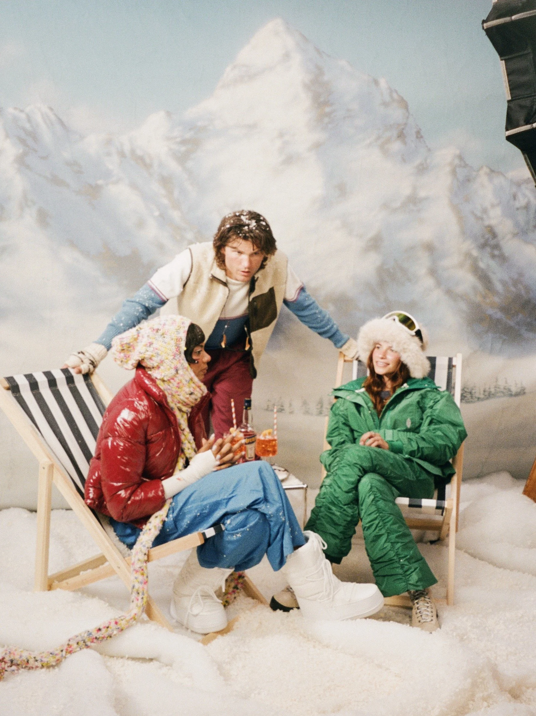 Five young adults dressed in winter clothes on a snowy outdoor set with a mountain backdrop. Two are seated on deck chairs, one in a red jacket and blue snow pants, another in green snow pants, and a third in a snowboarder outfit. Two others stand be