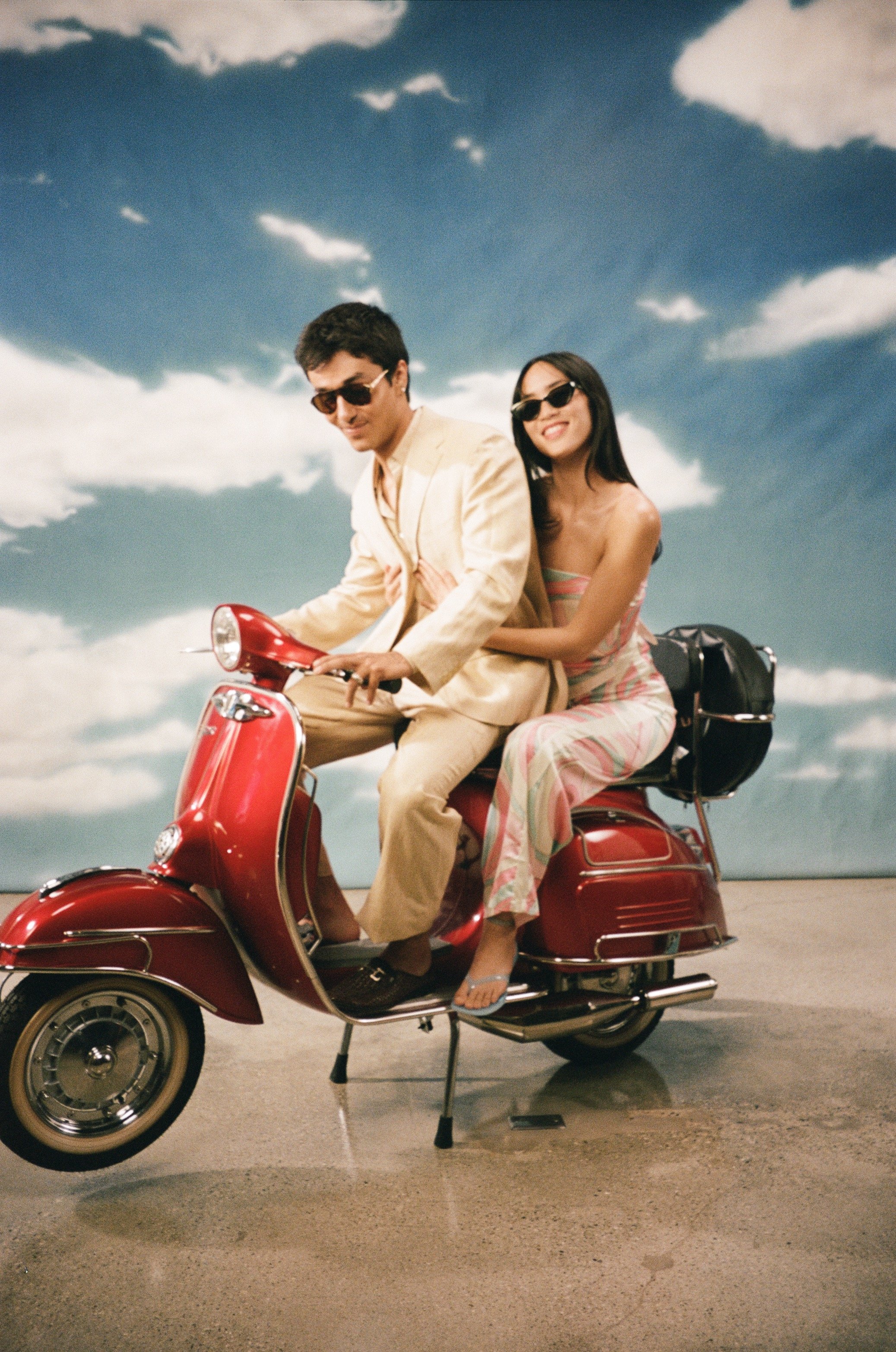 A man and woman riding a red scooter under a partly cloudy sky, both wearing sunglasses. The man is dressed in a light-colored suit and the woman in a colorful dress, smiling as they ride.