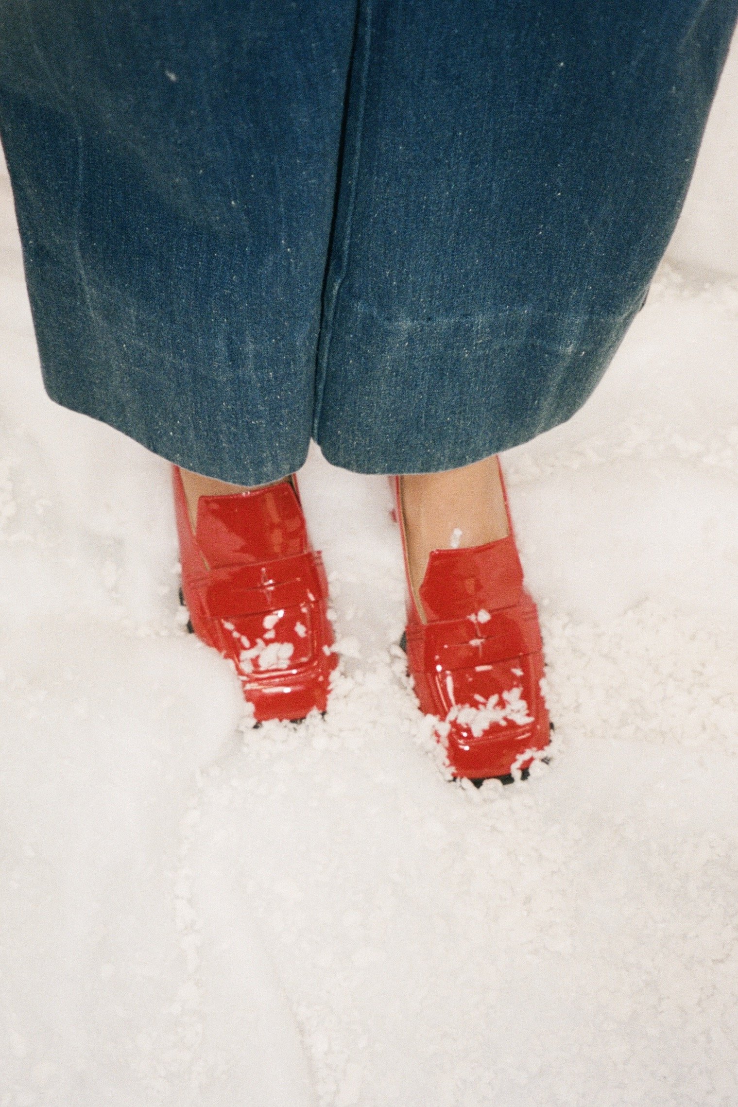 Person standing in snow wearing red rain boots, blue jeans, and a blue coat.