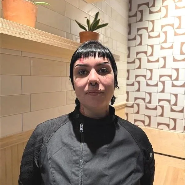 Smiling man with short curly hair and a beard, wearing a black jacket, sitting in front of a patterned wall.