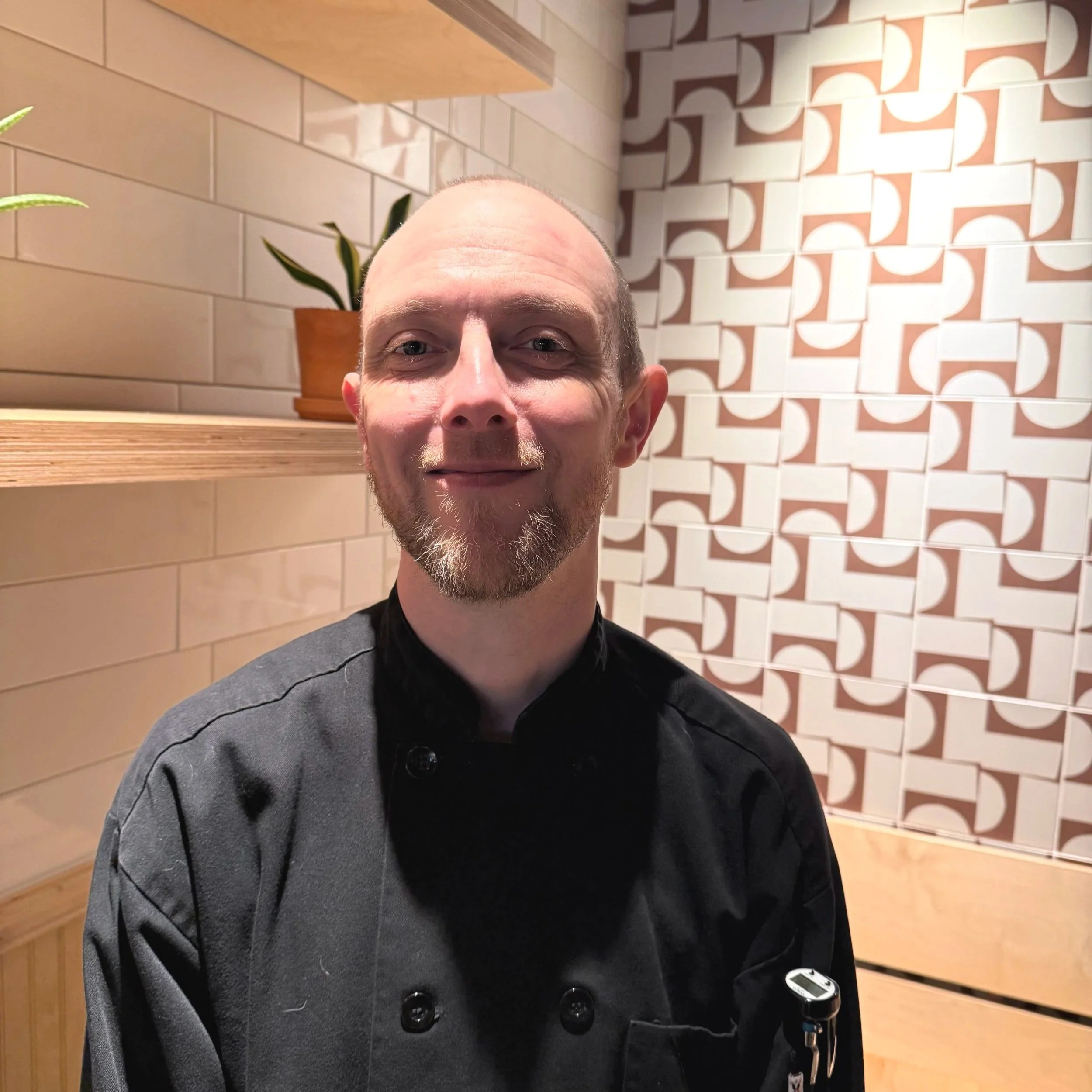 Smiling young woman with short curly hair, wearing a black hoodie, in a restaurant or cafe kitchen with two women in the background preparing food.