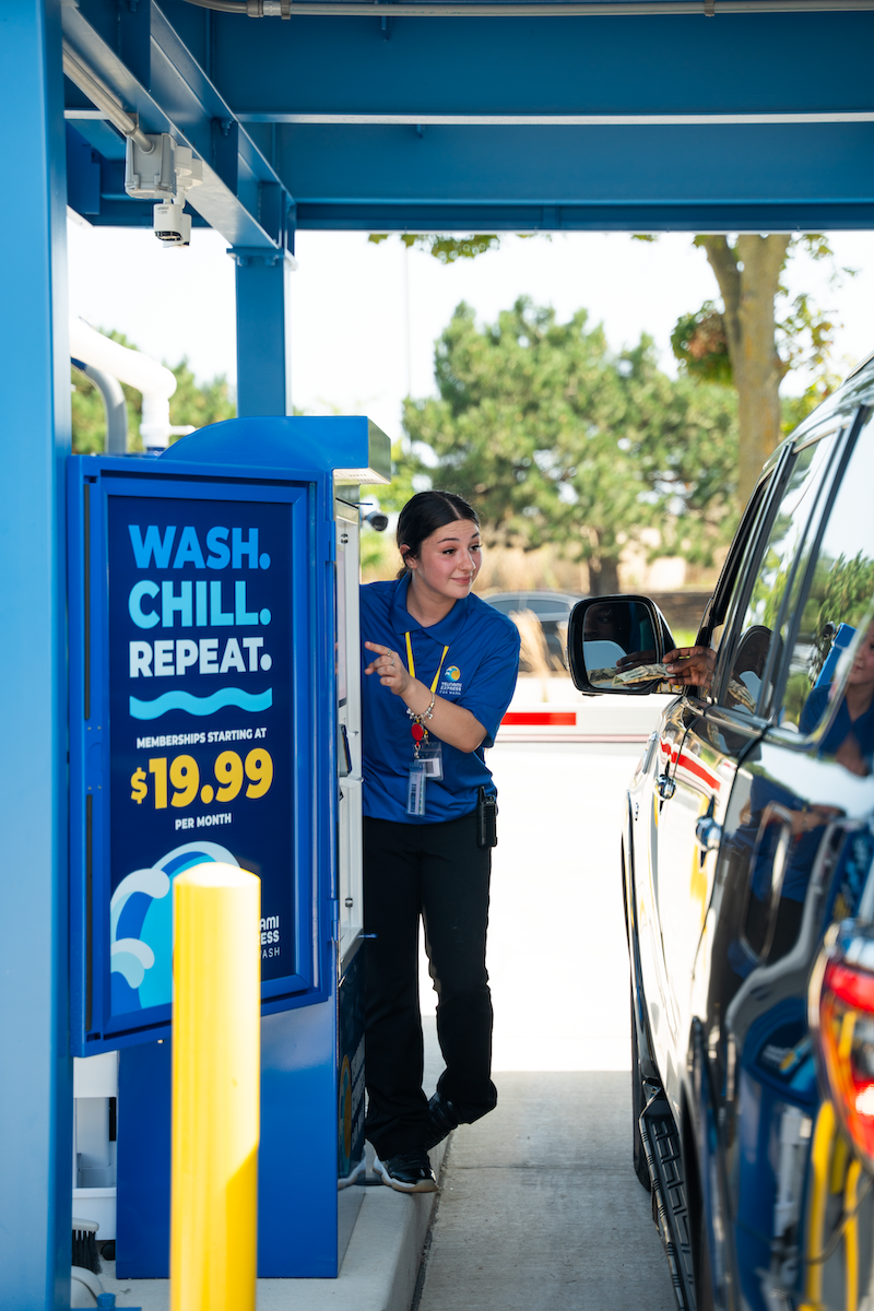A woman at a car wash booth talking to a person in a black vehicle through the window. The booth has a sign that says 'Wash. Chill. Repeat.' with membership starting at $19.99 per month.