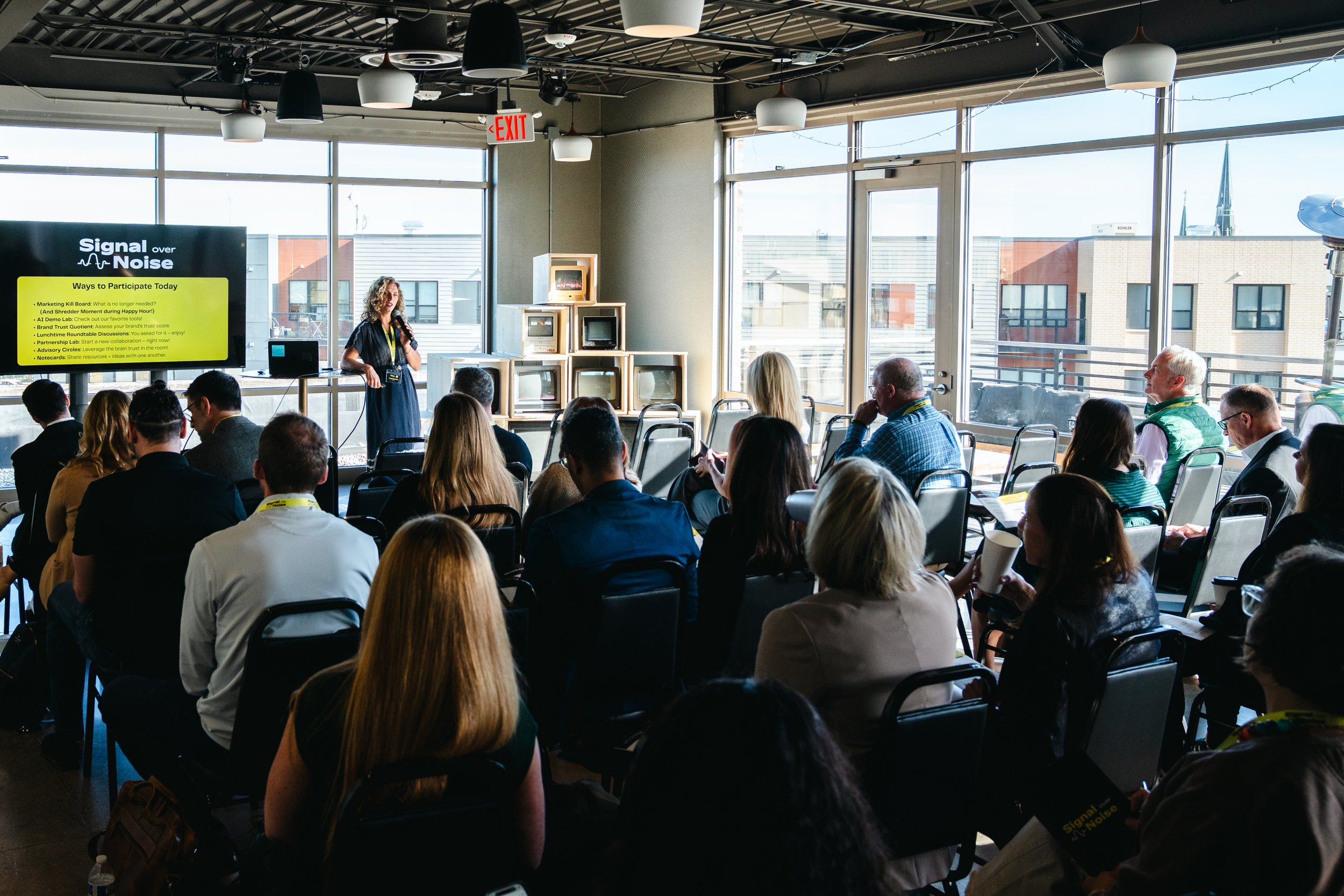 A woman is giving a presentation at a conference, standing in front of a large screen that displays the event schedule. The audience is seated and listening attentively in a bright room with large windows and modern decor.